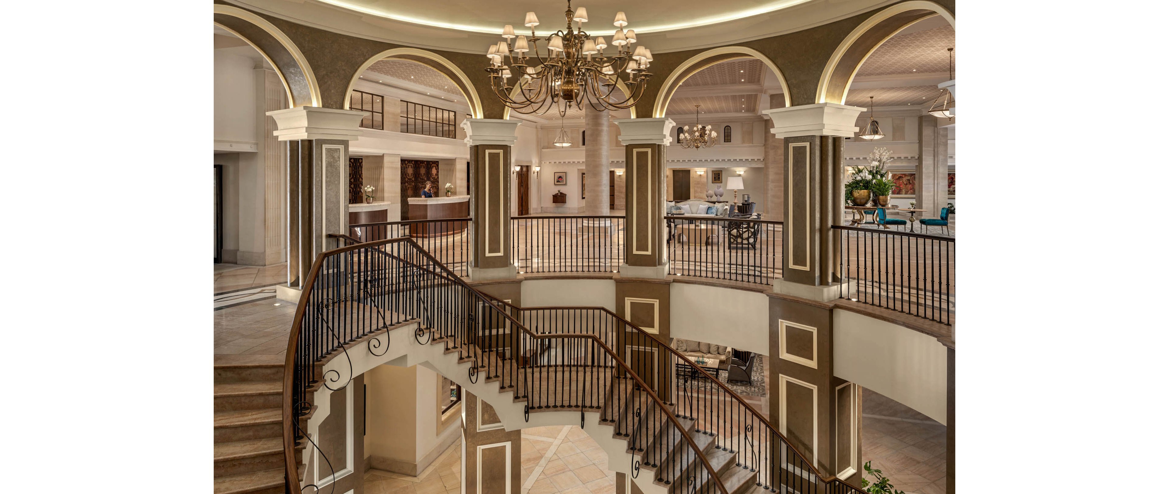 A luxurious hotel atrium features a sweeping curved staircase leading down to a lower level. The space is framed by tall columns, arched openings, and ornate railings that emphasize the grand architecture. A large chandelier hangs from a circular gold-toned ceiling detail, adding elegance and warmth. Soft ambient lighting and neutral stone flooring create a refined, welcoming atmosphere.