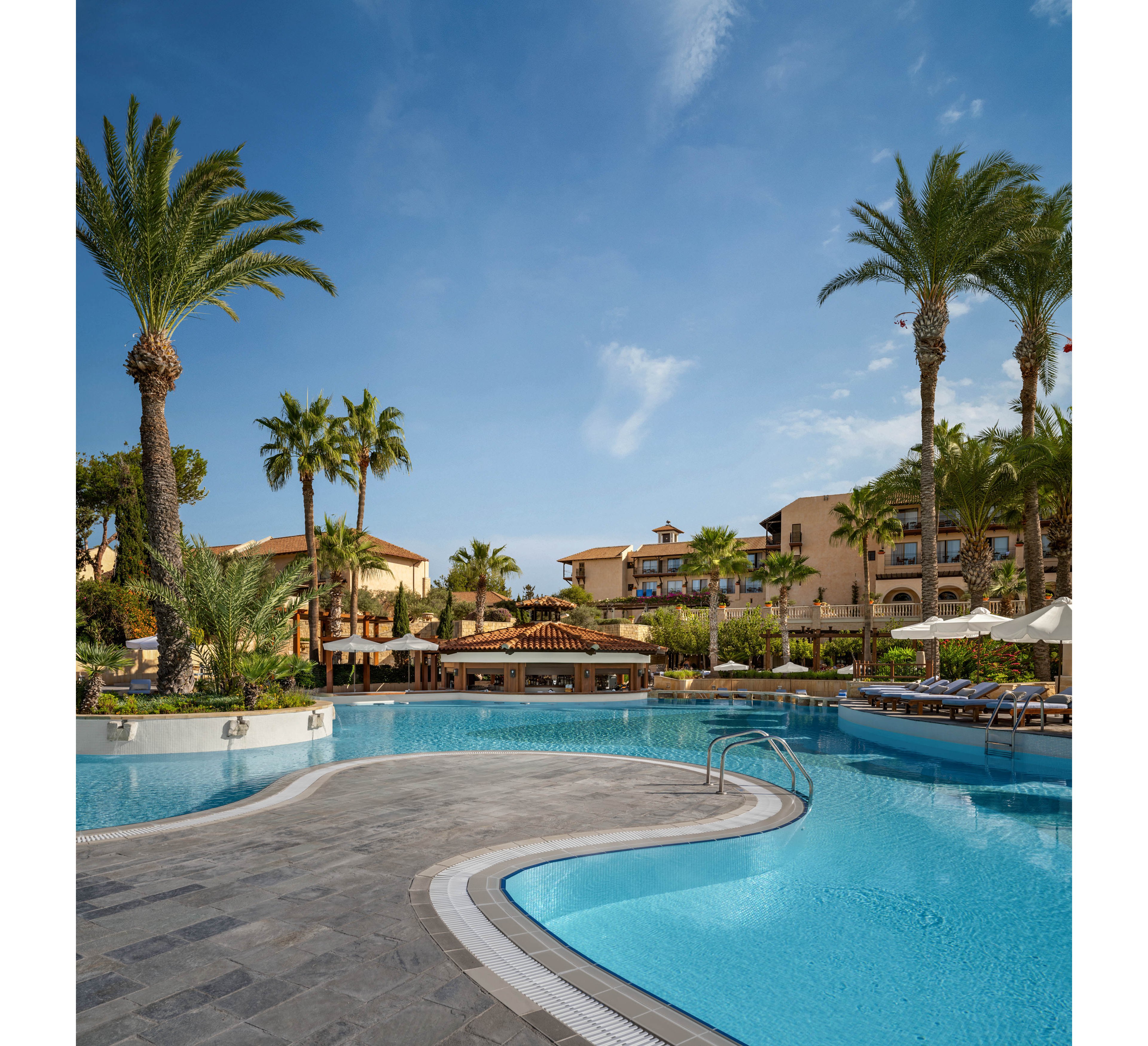 A large resort swimming pool curves gracefully under a clear blue sky. Tall palm trees and lush greenery surround the water, creating a tropical atmosphere. In the background, multi-story hotel buildings and a poolside bar add to the vacation setting. Sun loungers and umbrellas line the far side of the pool, suggesting a relaxing holiday environment.