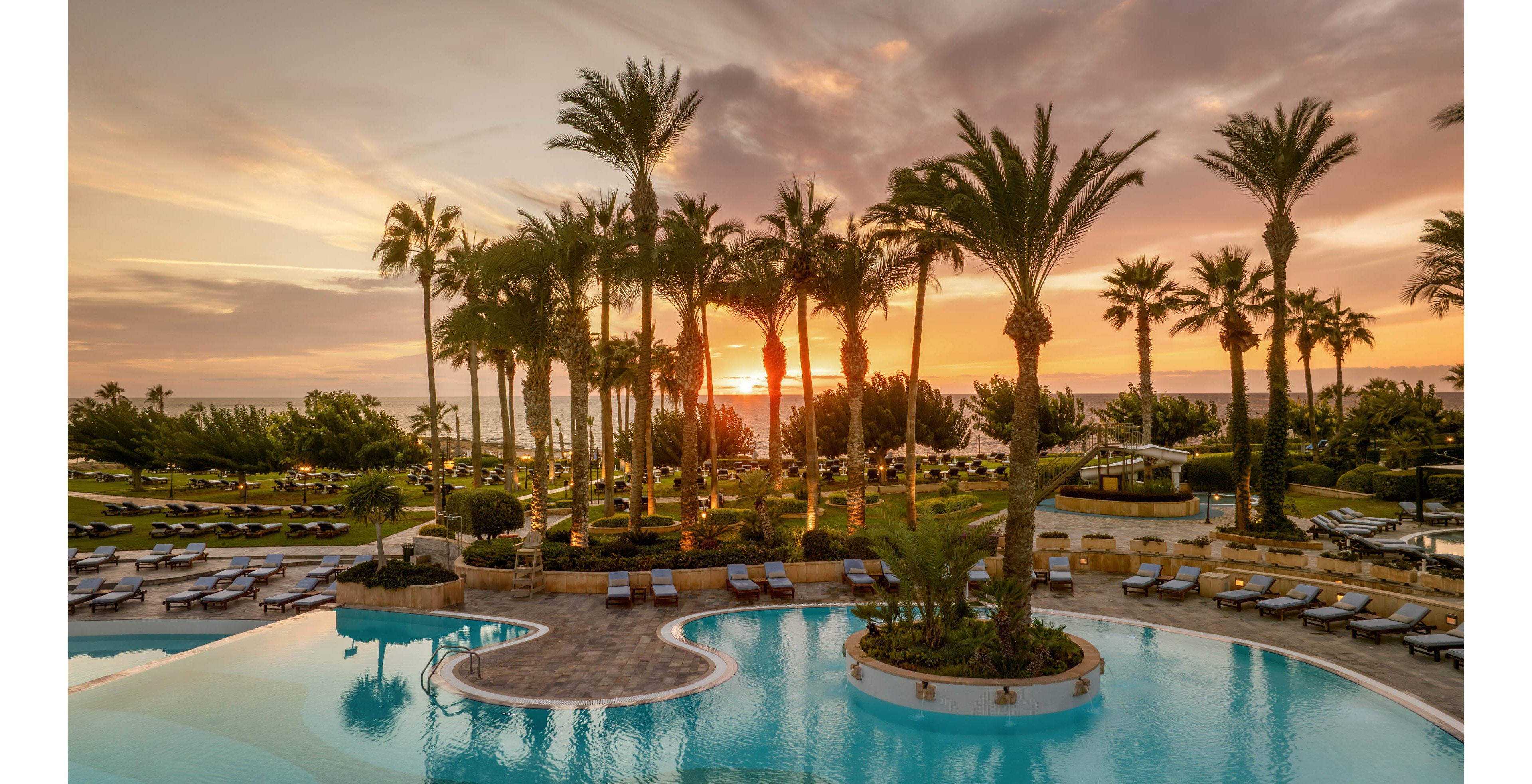 A large resort-style swimming pool curves gracefully in the foreground, surrounded by neatly arranged lounge chairs and umbrellas. Tall palm trees frame the scene as the sun sets over the distant sea, casting warm golden light across the water. The sky is filled with soft clouds in shades of orange, pink, and purple, reflecting on the calm pool surface. The overall atmosphere suggests a tranquil, upscale vacation destination by the coast.