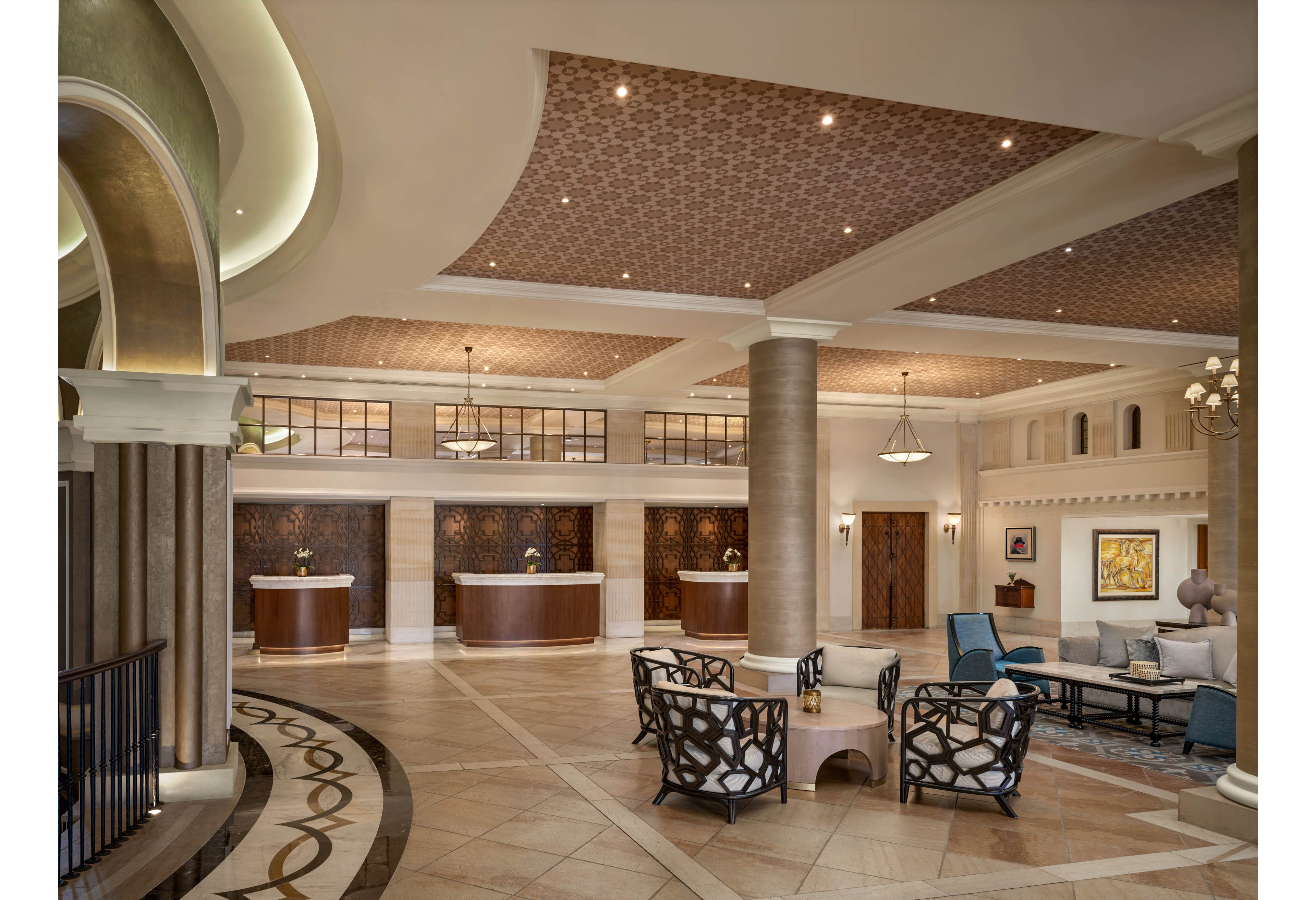 A large, elegant hotel lobby features multiple reception desks along the back wall. The space includes tall columns, patterned ceilings, and polished tile floors with decorative inlays. Comfortable seating areas with armchairs and coffee tables are arranged throughout the lobby. Warm lighting and framed artwork contribute to a sophisticated, welcoming atmosphere.