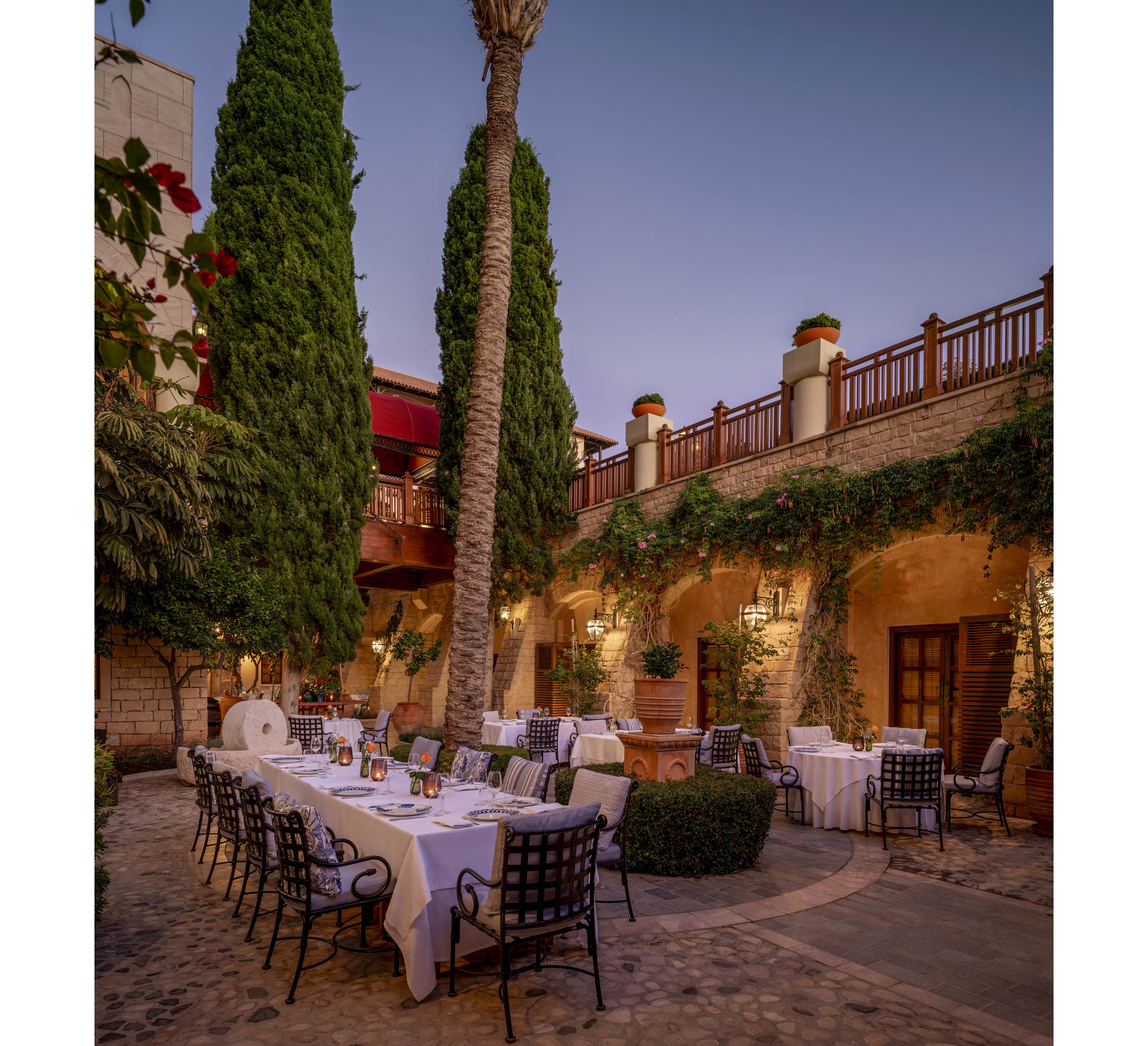 An elegant outdoor dining area is set in a stone courtyard at dusk, with long white-clothed tables and black metal chairs arranged for guests. Warm ambient lighting glows from wall sconces and lanterns, highlighting the arches and textured stone walls. Tall palm trees and lush climbing vines frame the space, while a wooden balcony and railings overlook the courtyard. The tranquil atmosphere suggests a refined restaurant or event setting in a Mediterranean or historic-style venue.