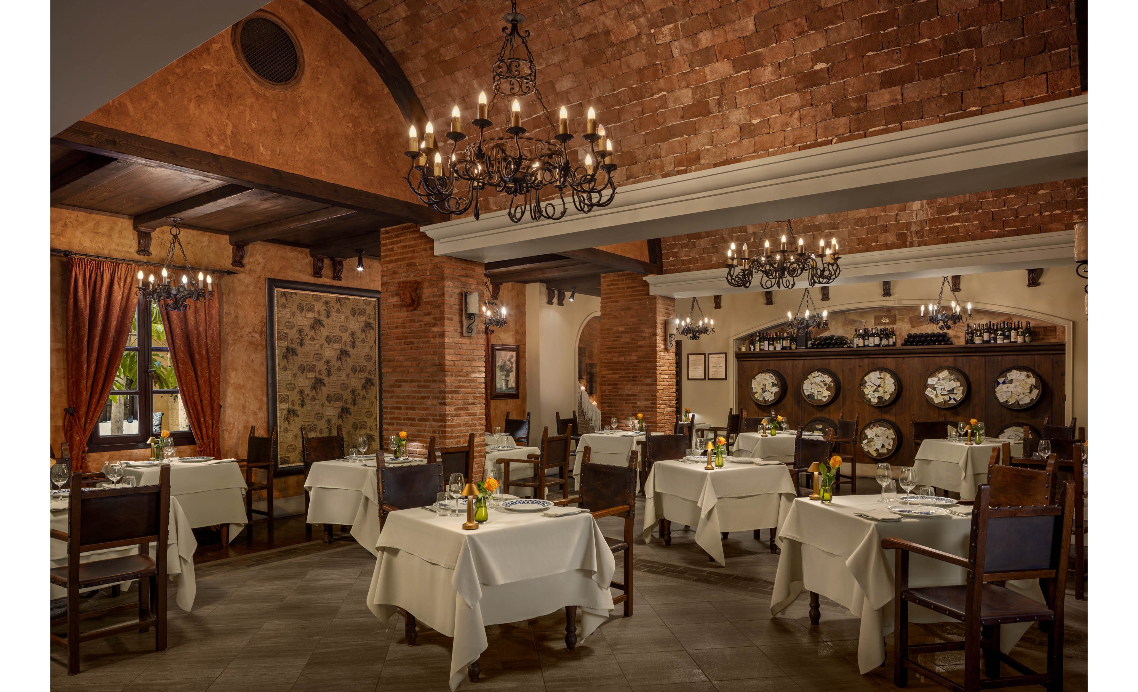 A warmly lit restaurant dining room features neatly arranged tables covered with white tablecloths. Dark wooden chairs and chandeliers complement the rustic brick walls and arched ceiling. Decorative plates line a recessed wall, while curtains and framed artwork add to the cozy ambiance. The space appears ready for guests, with place settings and small floral centerpieces on each table.