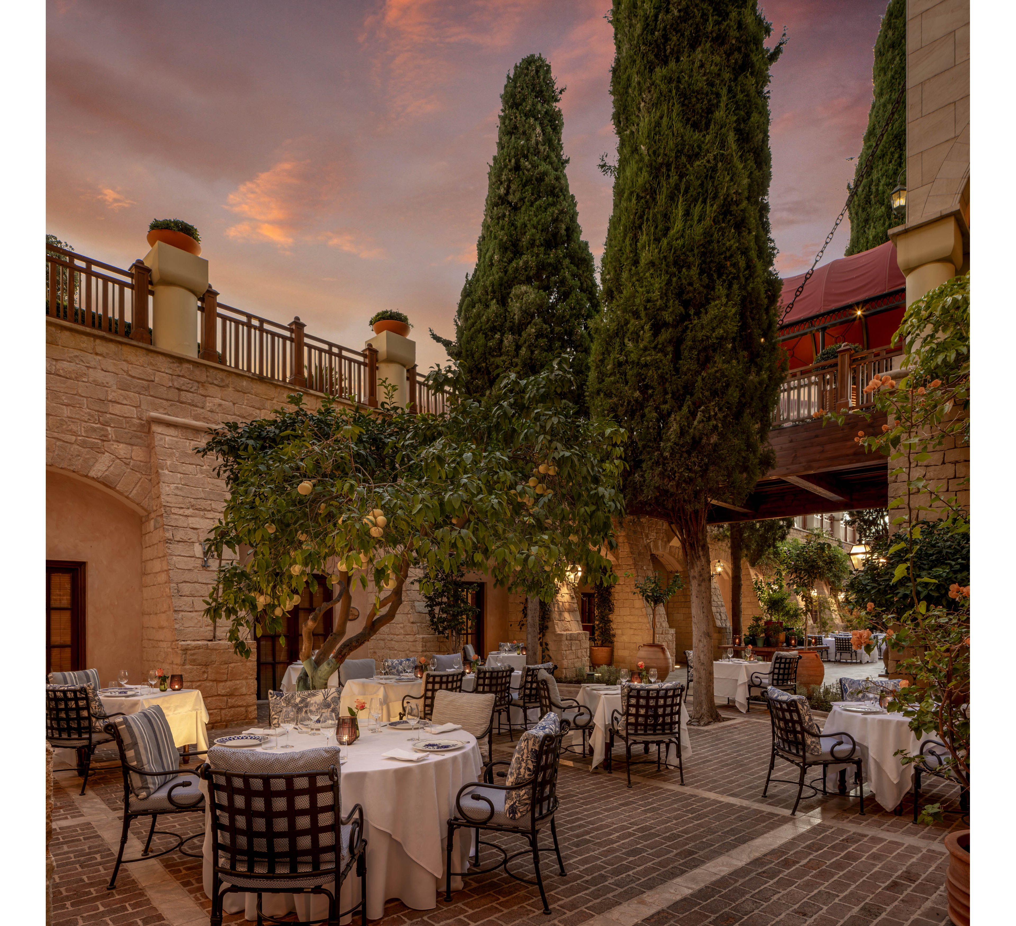 An elegant outdoor restaurant is set within a stone courtyard surrounded by tall cypress trees. White tablecloths and cushioned metal chairs are arranged neatly around the patio, ready for evening diners. Warm string lights and subtle lanterns create a cozy ambiance as the sky glows with soft sunset colors. The scene feels intimate and romantic, blending natural greenery with refined architectural details.