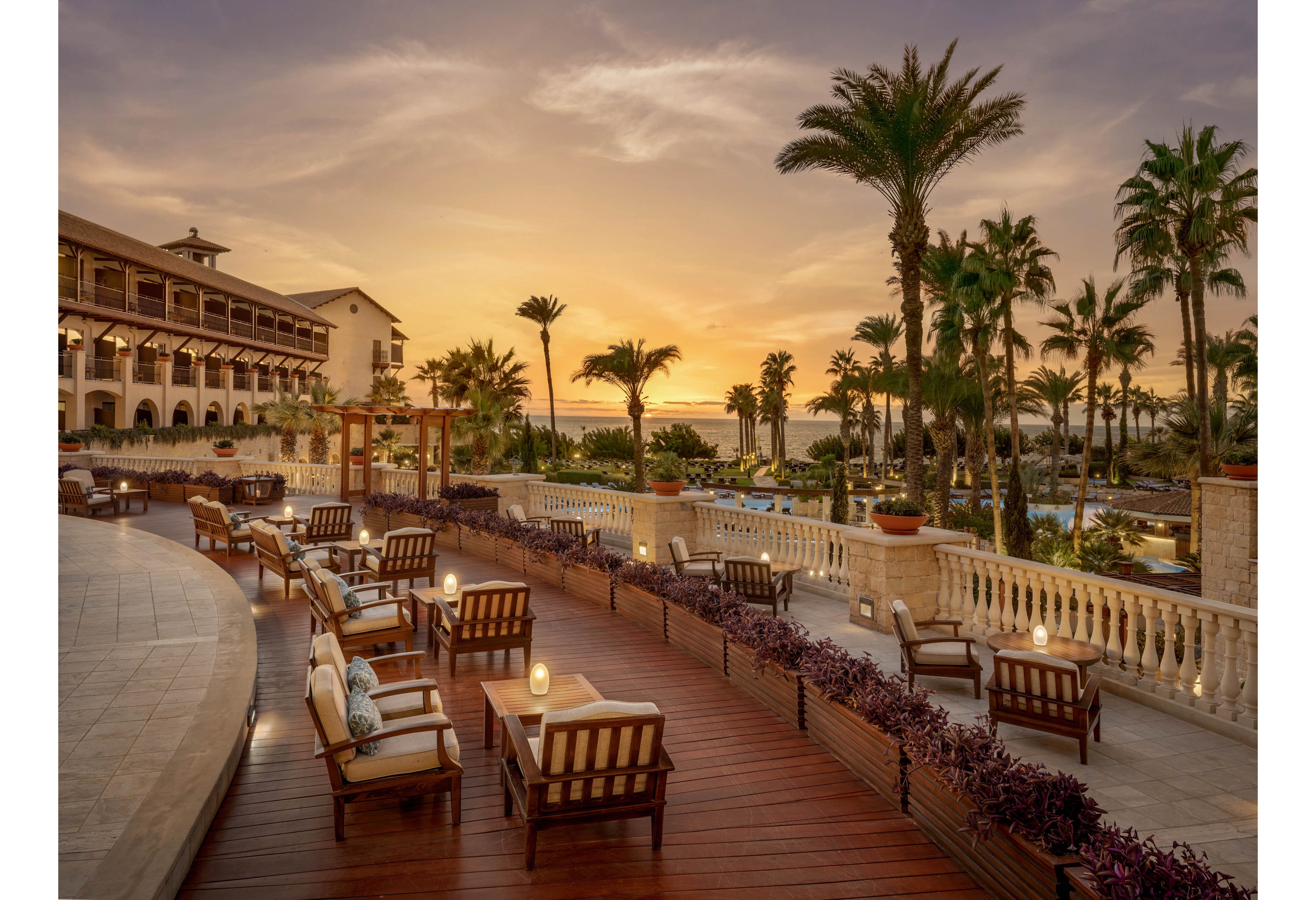 An elegant resort terrace overlooks a lush tropical landscape at sunset. Wooden decking is furnished with cushioned lounge chairs and small tables, some topped with glowing candles. Palm trees frame the view toward a distant pool area and the ocean horizon. Warm golden light and a pastel sky create a tranquil, romantic atmosphere ideal for evening relaxation.