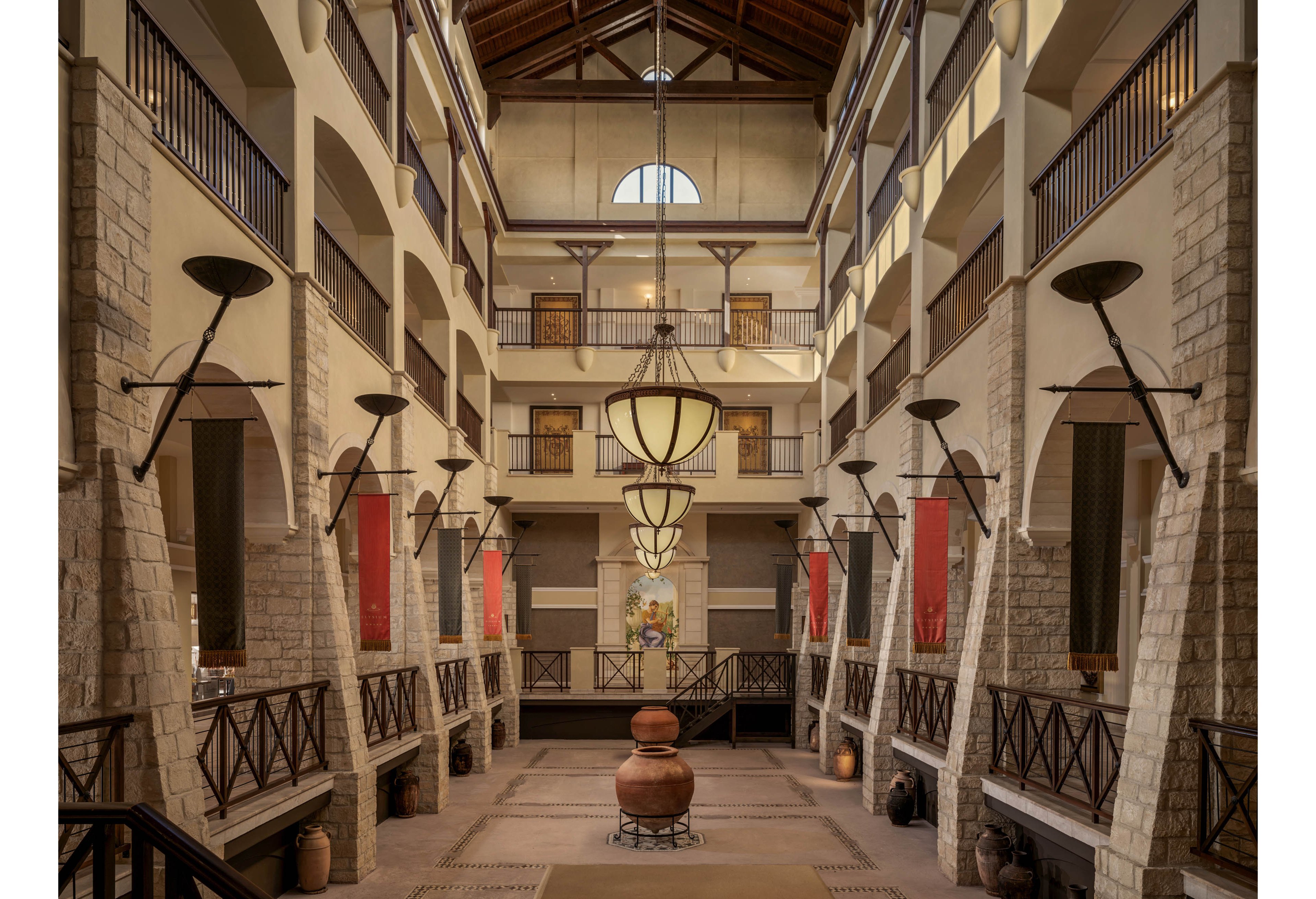 A spacious hotel atrium features tall stone columns and multiple balconies overlooking the central hall. Warm pendant lights hang from the high wooden ceiling, illuminating the symmetrical architecture. Large clay pots and decorative wall banners add rustic character to the space. The overall scene feels inviting and elegant, with a balanced, symmetrical composition.