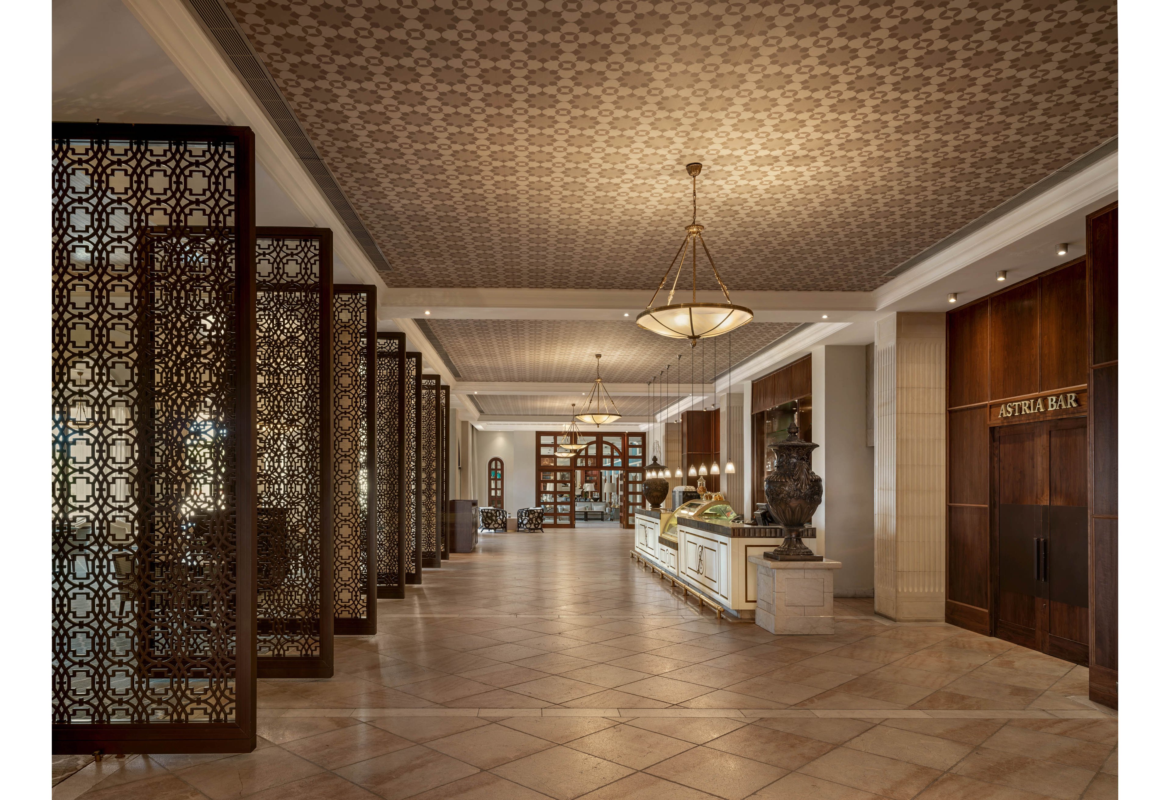 A spacious hotel lobby features intricate lattice partitions leading toward a distant seating area. Warm lighting from large chandeliers highlights the patterned ceiling and tiled floor. A central counter displays decorative objects and vases, adding to the refined atmosphere. On the right, a wooden entrance labeled "ASTRIA BAR" suggests an adjoining bar area within the hotel.