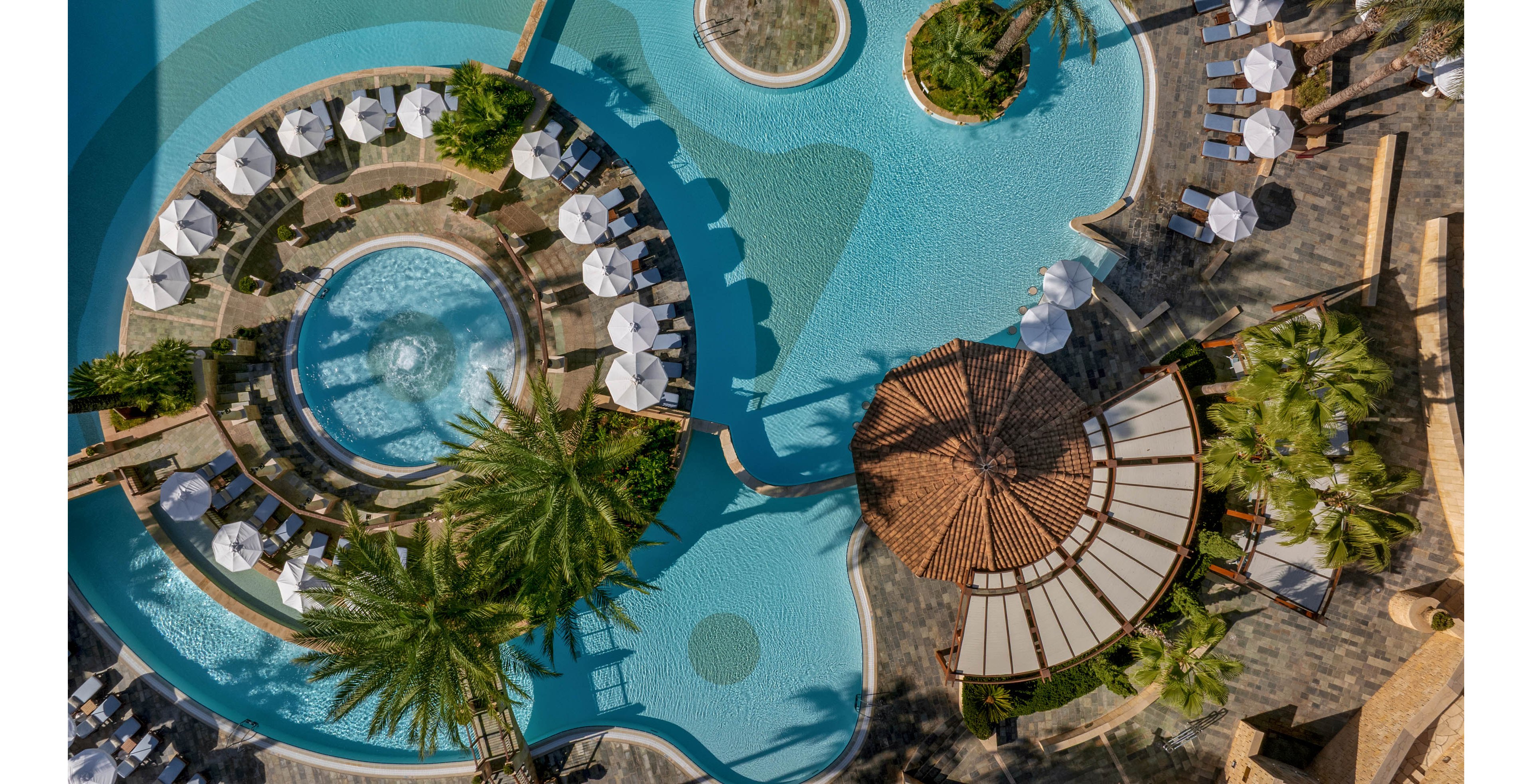 An aerial view showcases a luxurious resort pool complex with curved turquoise swimming pools. Circular islands with palm trees and sun loungers are surrounded by neatly arranged white umbrellas. A shaded pavilion with a terracotta roof sits beside the water, adding architectural interest. The bright sunlight and clear blue water create a relaxing, vacation-like atmosphere.