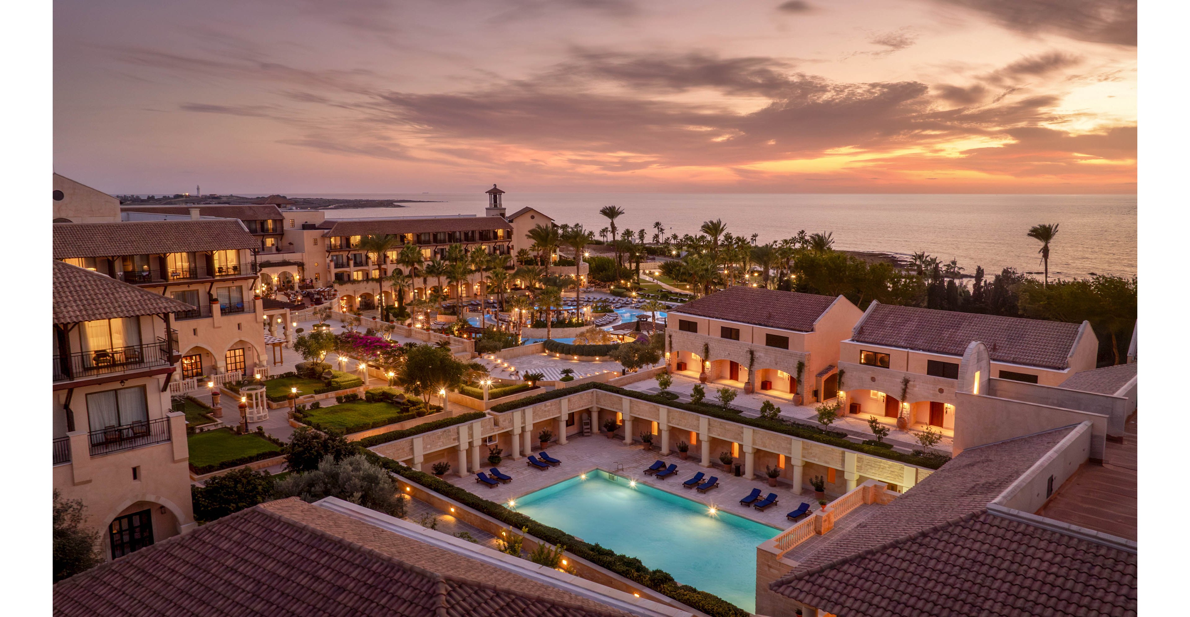 A large luxury resort complex is captured at sunset overlooking the sea. Multiple illuminated swimming pools and palm-lined walkways create a vibrant yet relaxed atmosphere. The architecture features warm-toned buildings with tiled roofs surrounding central courtyards. Soft evening lighting and a colorful sky enhance the tranquil coastal vacation setting.