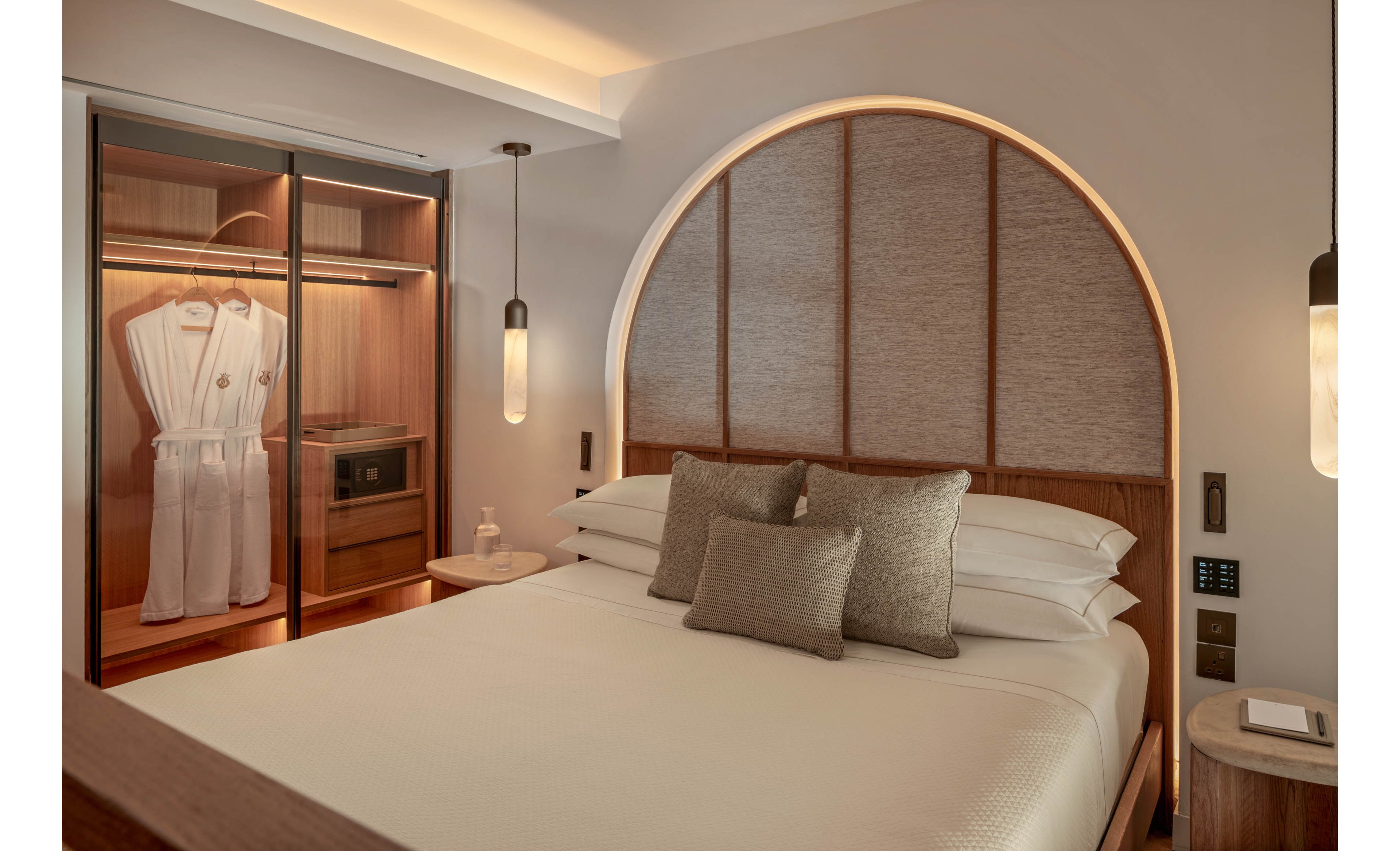 A contemporary hotel bedroom features a neatly made bed with multiple neutral-toned pillows and a softly lit arched headboard. To the left, an open wardrobe reveals two hanging bathrobes, shelves, and a built-in safe or microwave. Warm wooden finishes and soft ambient lighting create a calm, inviting atmosphere. Minimal decor and clean lines emphasize a modern, luxurious design.