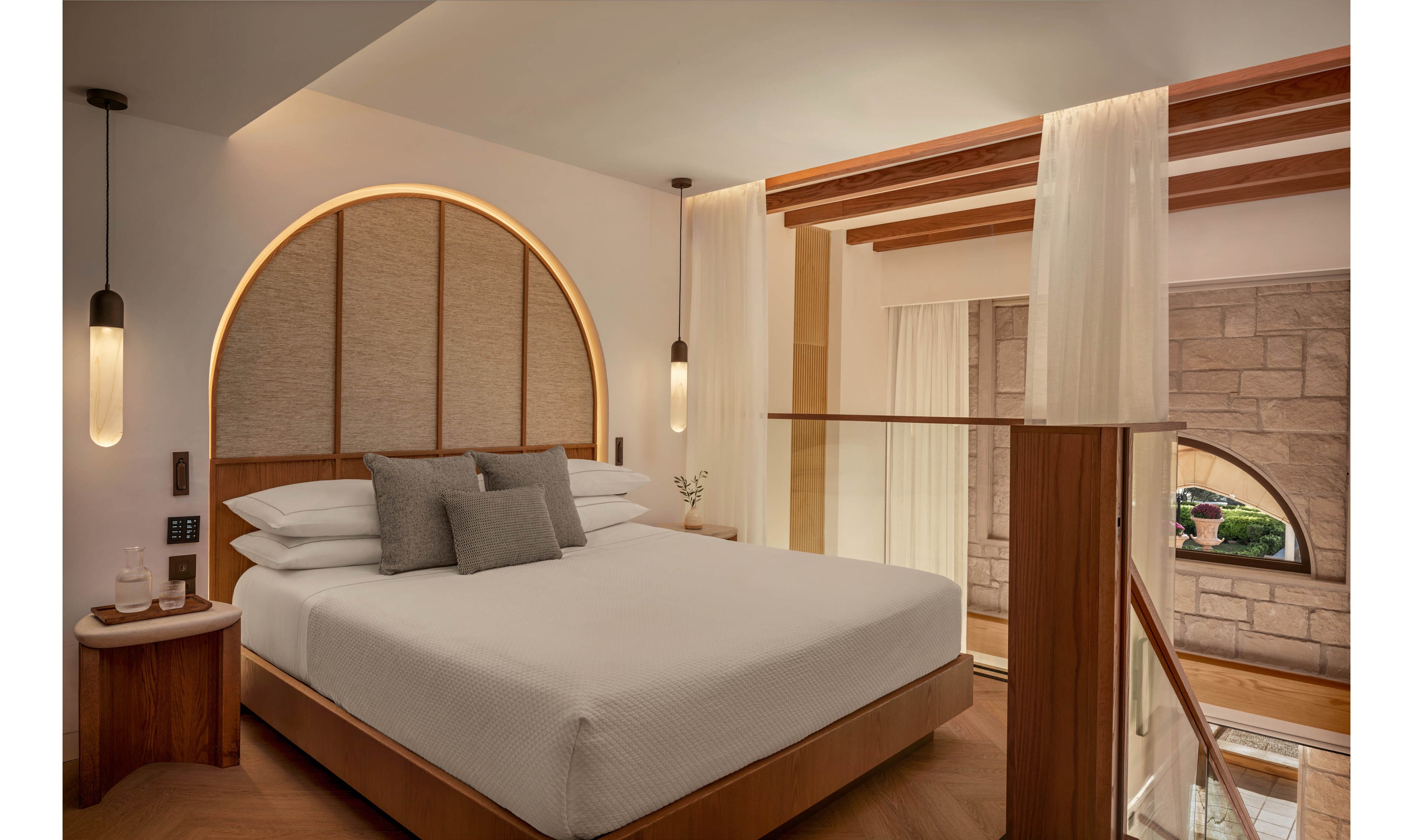 A contemporary hotel bedroom features a neatly made bed with white linens and gray accent pillows. The room showcases a large arched headboard with integrated warm lighting and matching wooden furniture. Sheer curtains separate the sleeping area from a lower level, where a window reveals a stone wall and greenery outside. Pendant lights and soft neutral tones create a calm, inviting atmosphere.