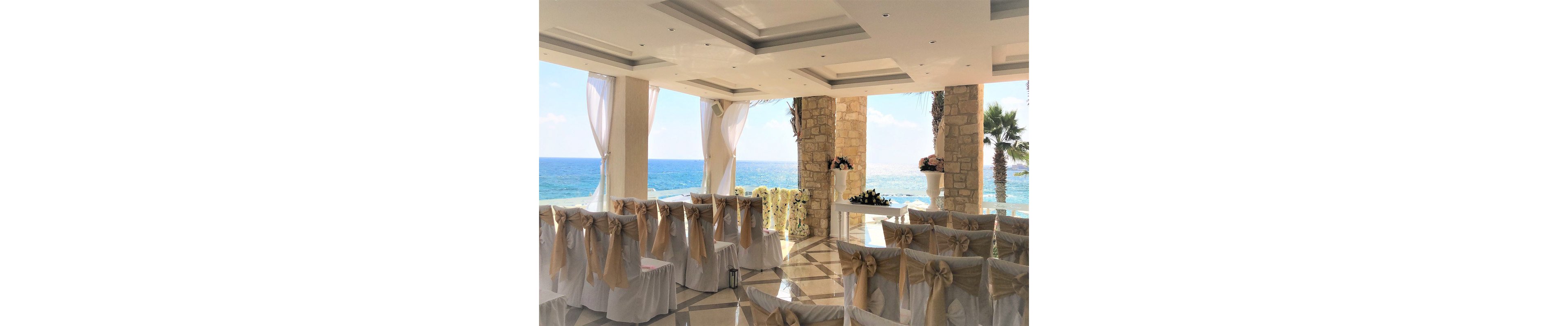 Rows of white chairs with beige bows are arranged for a wedding ceremony in an open-air venue overlooking the sea. Sheer white curtains frame the bright blue ocean and sky, creating a romantic coastal backdrop. Stone columns and floral arrangements add a touch of sophistication to the space. Sunlight reflects off the polished floor, enhancing the airy and celebratory atmosphere.