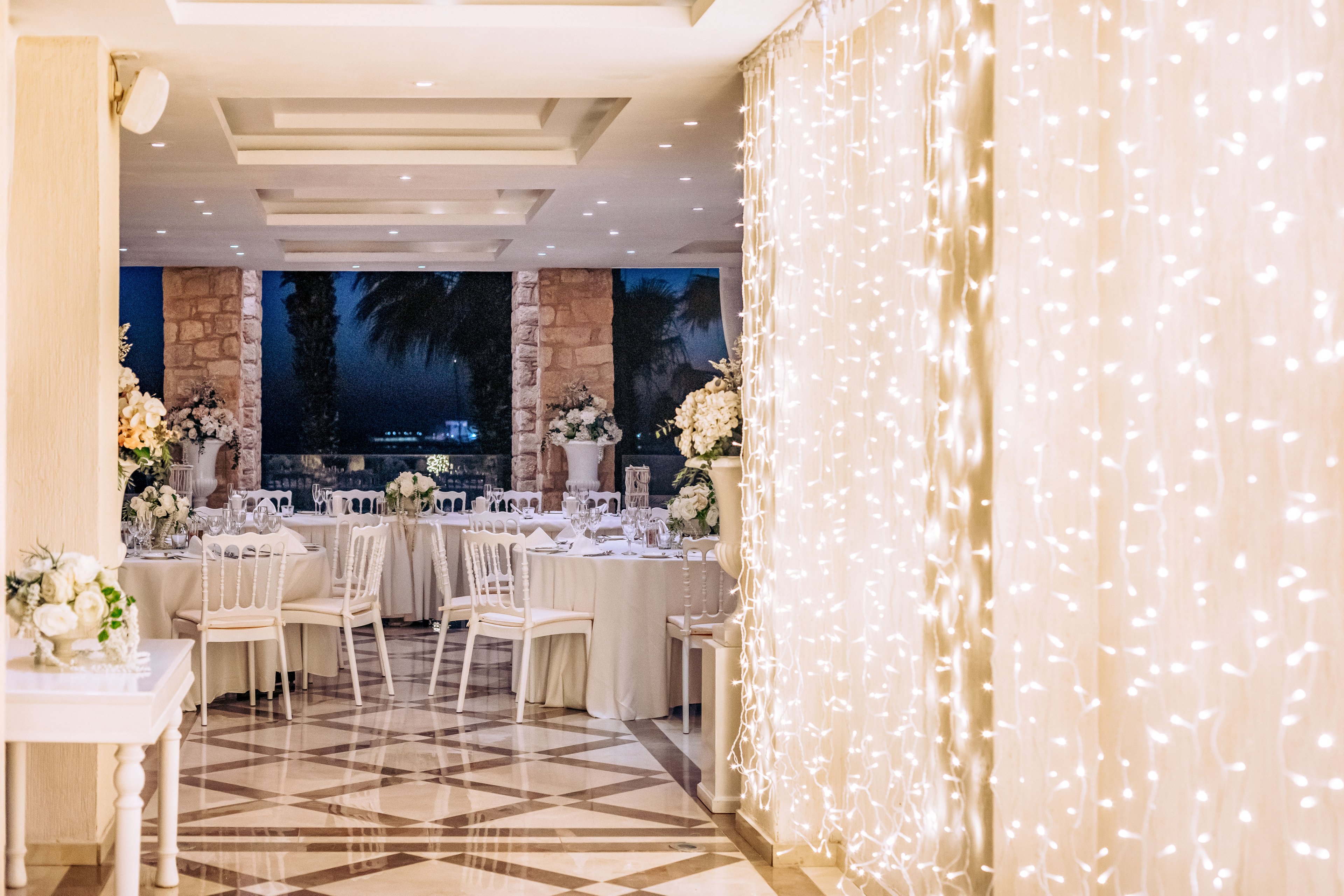 A beautifully decorated indoor wedding reception space features round tables with white linens and floral centerpieces. White chairs surround the tables, arranged near large stone columns that open to a nighttime outdoor view. A curtain of warm fairy lights glows along one side of the room, reflecting softly on the polished patterned floor. The overall atmosphere is romantic and sophisticated, with soft lighting and neutral tones creating an intimate celebration setting.