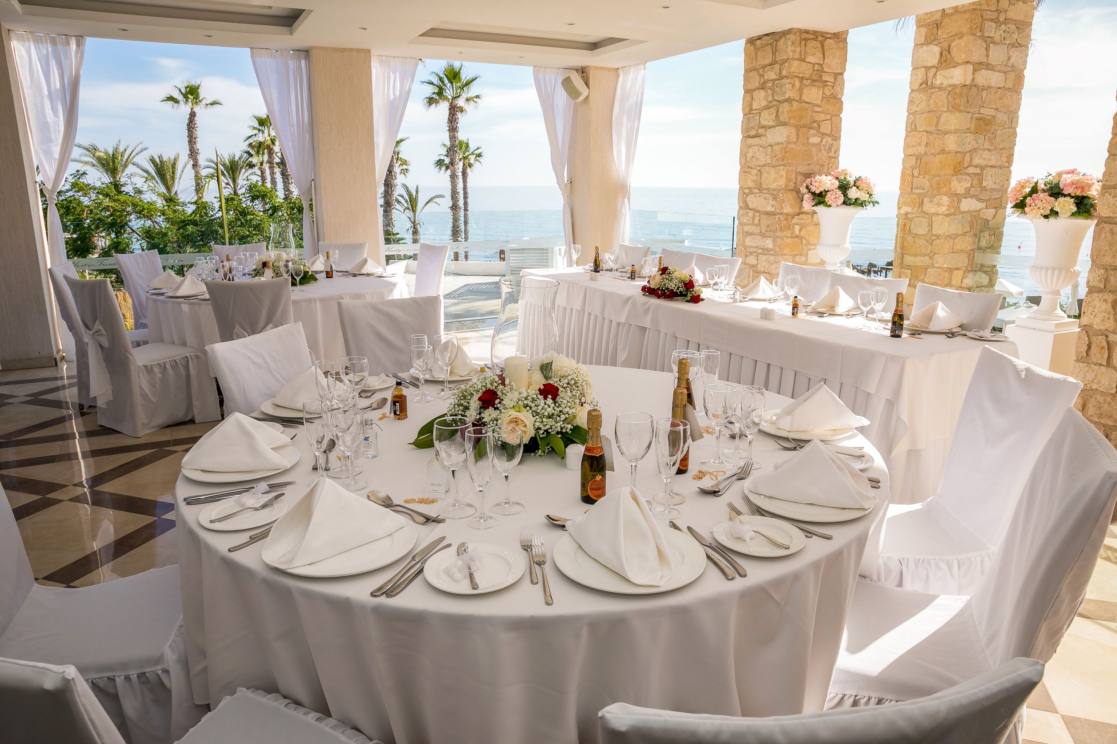 Round banquet tables are beautifully arranged for a formal event in a bright seaside venue. White tablecloths, folded napkins, and neatly placed glassware and cutlery create a refined dining setting. Floral centerpieces with red and white flowers add color to the neutral decor. Large windows and open curtains reveal palm trees and an ocean view, enhancing the airy, romantic atmosphere.