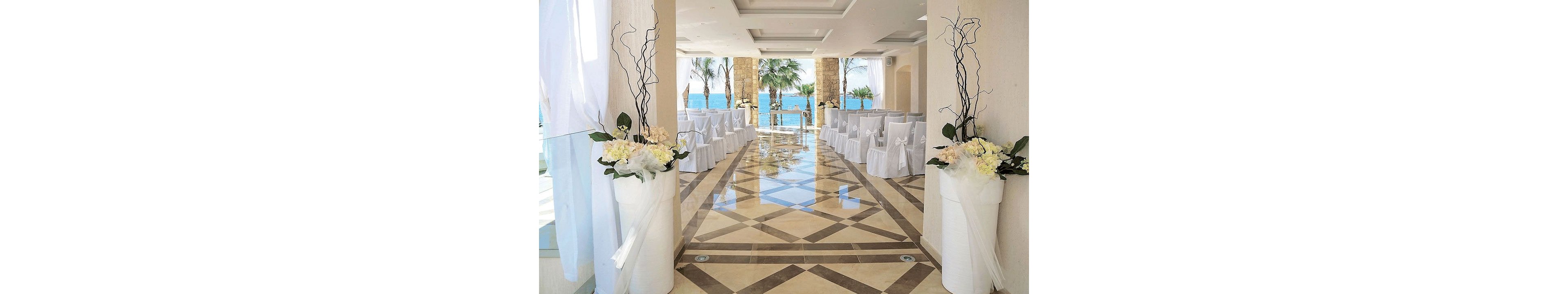 A bright, elegant indoor aisle is arranged for a wedding ceremony overlooking the sea. White chairs with bows line both sides of the polished geometric-tiled floor, leading toward an open view of palm trees and blue water. Tall white floral arrangements with branches frame the entrance, complemented by sheer white drapes and soft natural light. The scene conveys a sophisticated, romantic atmosphere in a coastal venue.