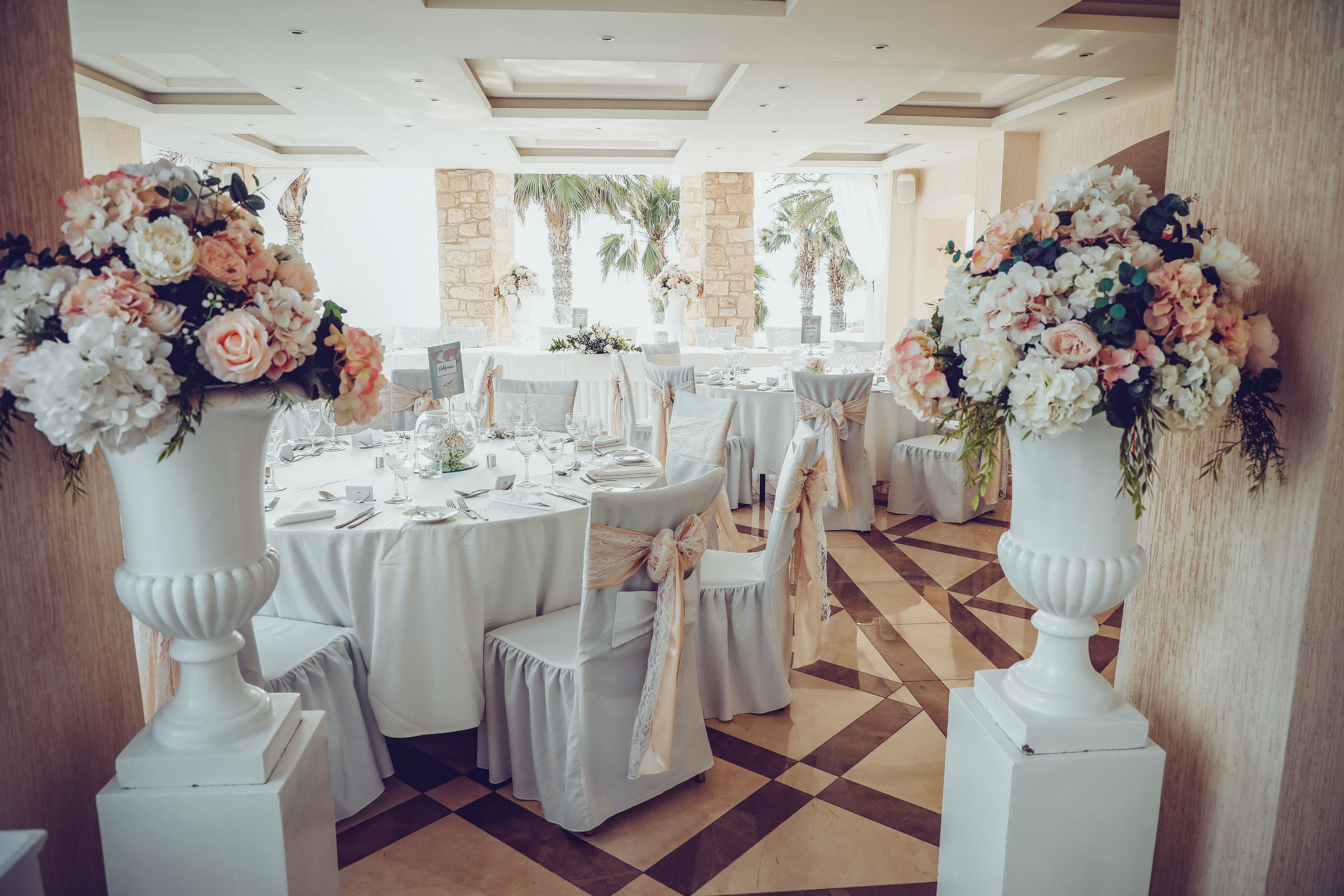 A beautifully decorated indoor reception hall features round tables draped in white linens and tied with soft beige chair sashes. Large white vases overflowing with pastel flowers frame the entrance, leading toward the center of the room. Glassware, plates, and subtle table decor are neatly arranged, suggesting a formal event or wedding celebration. Sunlight filters through large openings, revealing palm trees and stone columns outside, adding a warm, romantic atmosphere.
