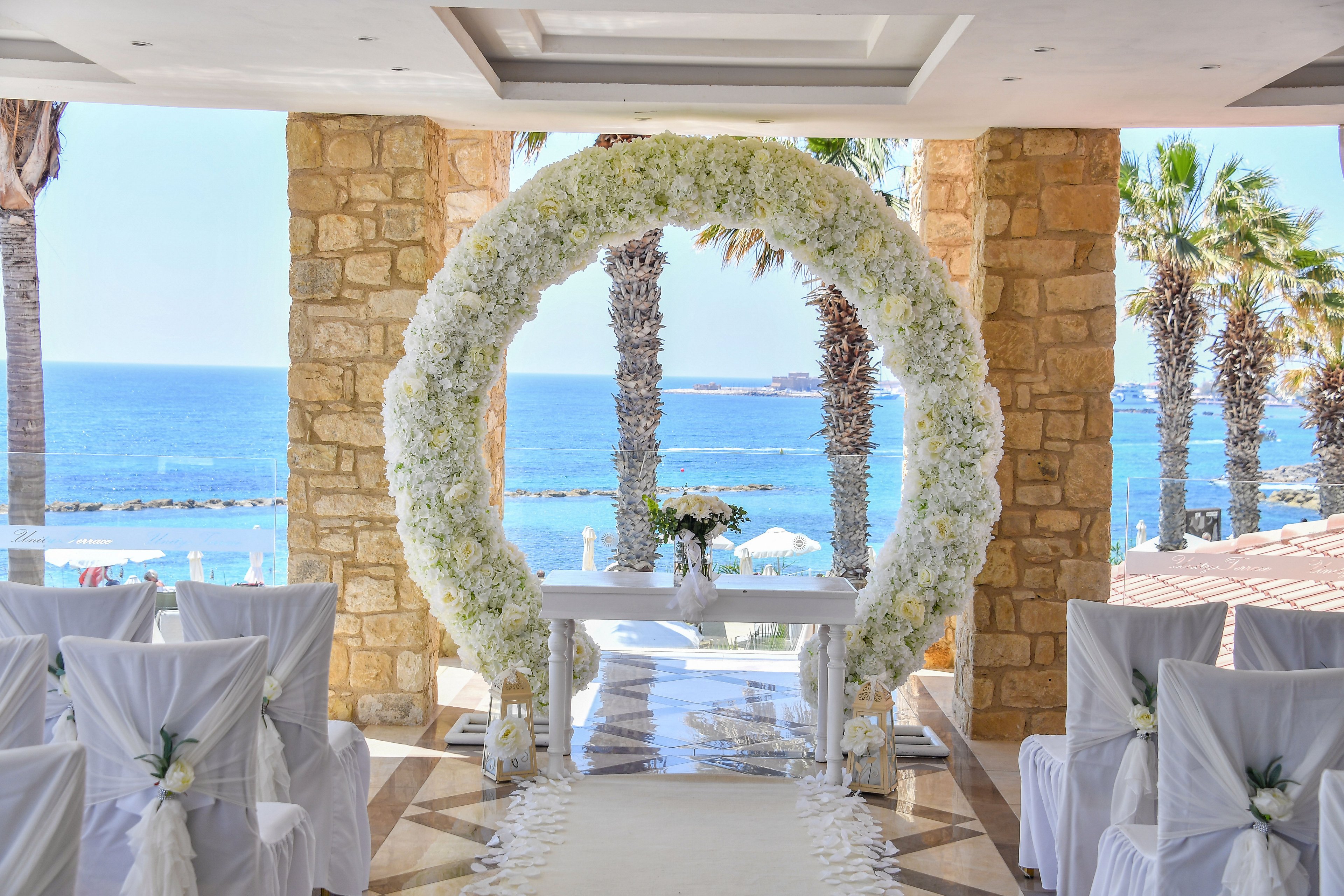 An elegant wedding ceremony setup overlooks a bright blue sea, framed by stone columns and palm trees. A large circular arch covered in white flowers surrounds a simple white altar table with a floral arrangement. Rows of chairs draped in white fabric line the aisle, each decorated with small floral accents. Sunlight reflects off the water, creating a romantic and serene coastal atmosphere.