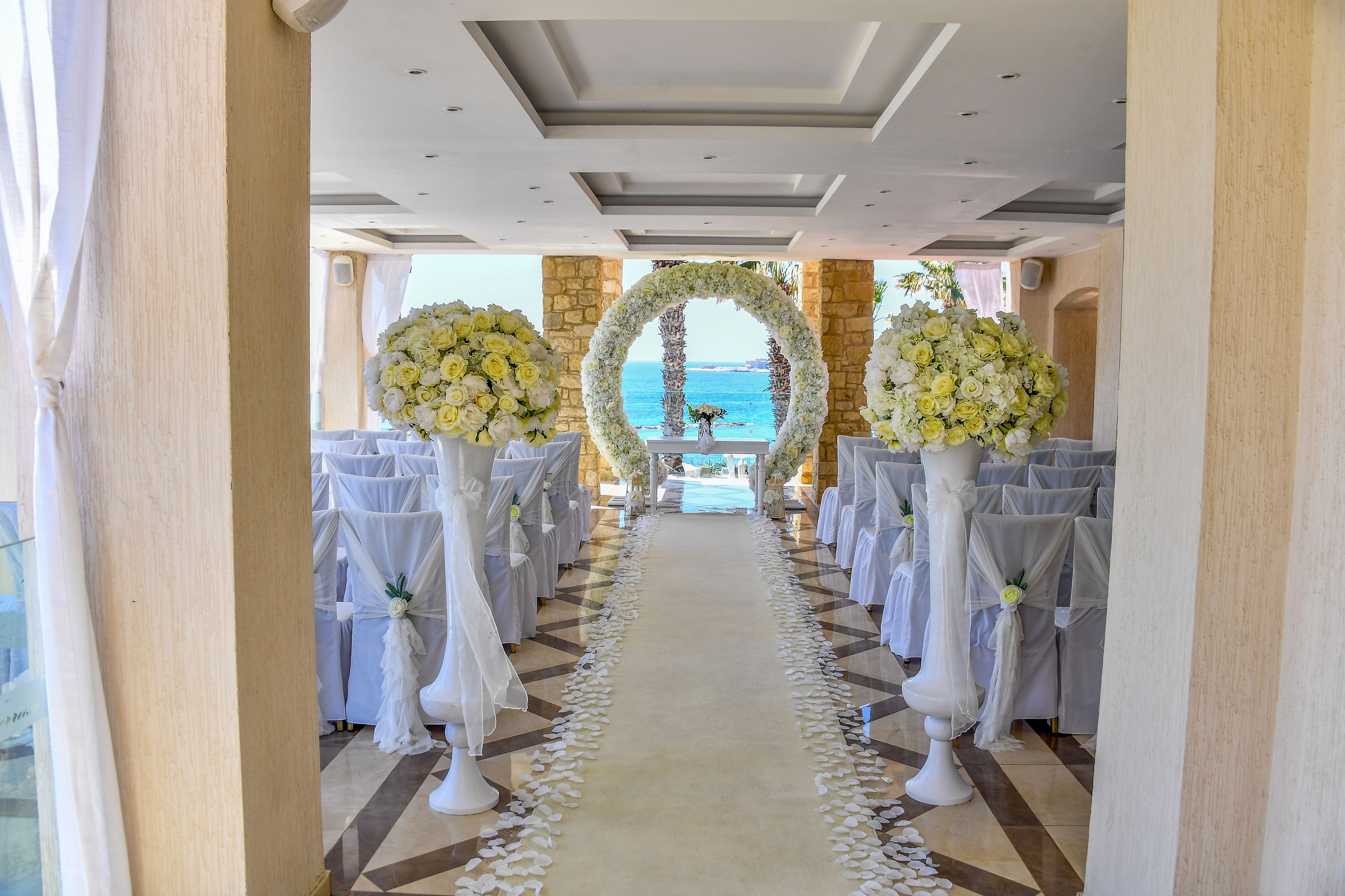 A beautifully decorated wedding ceremony aisle leads toward a floral arch framing a view of the sea. White chairs are neatly arranged on both sides, covered with light fabric and accented by small floral arrangements. Tall vases overflowing with white and yellow flowers stand at the entrance of the aisle, while petals are scattered along the floor. The bright natural light and ocean backdrop create a romantic, airy atmosphere.