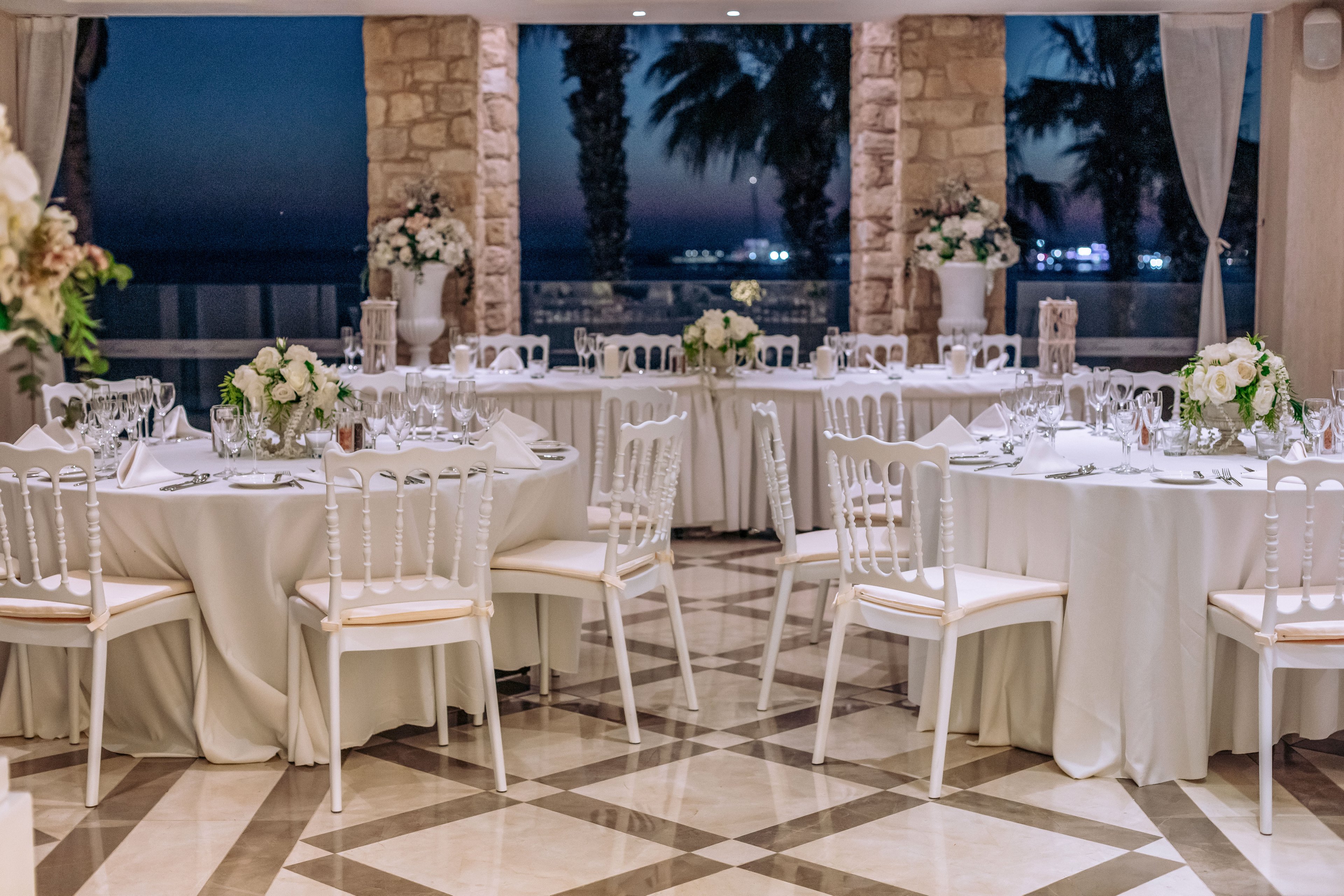 A beautifully decorated reception hall features round tables covered in white linens and surrounded by white chairs. Each table is set with glassware, plates, and floral centerpieces in soft white and green tones. Large stone columns frame windows that open to a twilight seaside view with palm trees and distant lights. The polished floor with geometric patterns adds to the sophisticated and romantic atmosphere.