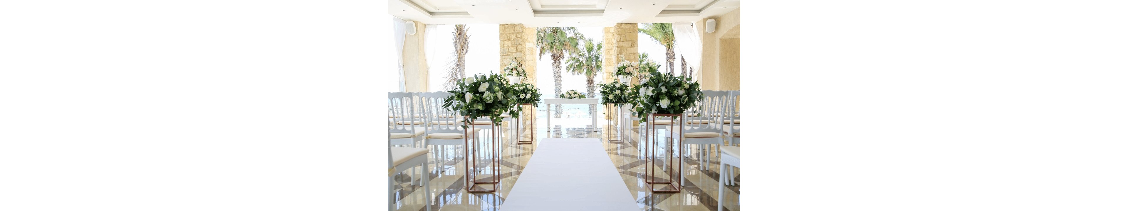 Bright wedding ceremony setup with a white aisle leading to an altar framed by large windows and palm trees. Rows of white chairs line both sides of the aisle, creating a symmetrical and formal arrangement. Tall stands hold lush green and white floral arrangements, adding a fresh, romantic touch. Sunlight reflects off the polished floor, enhancing the airy, coastal atmosphere of the venue.
