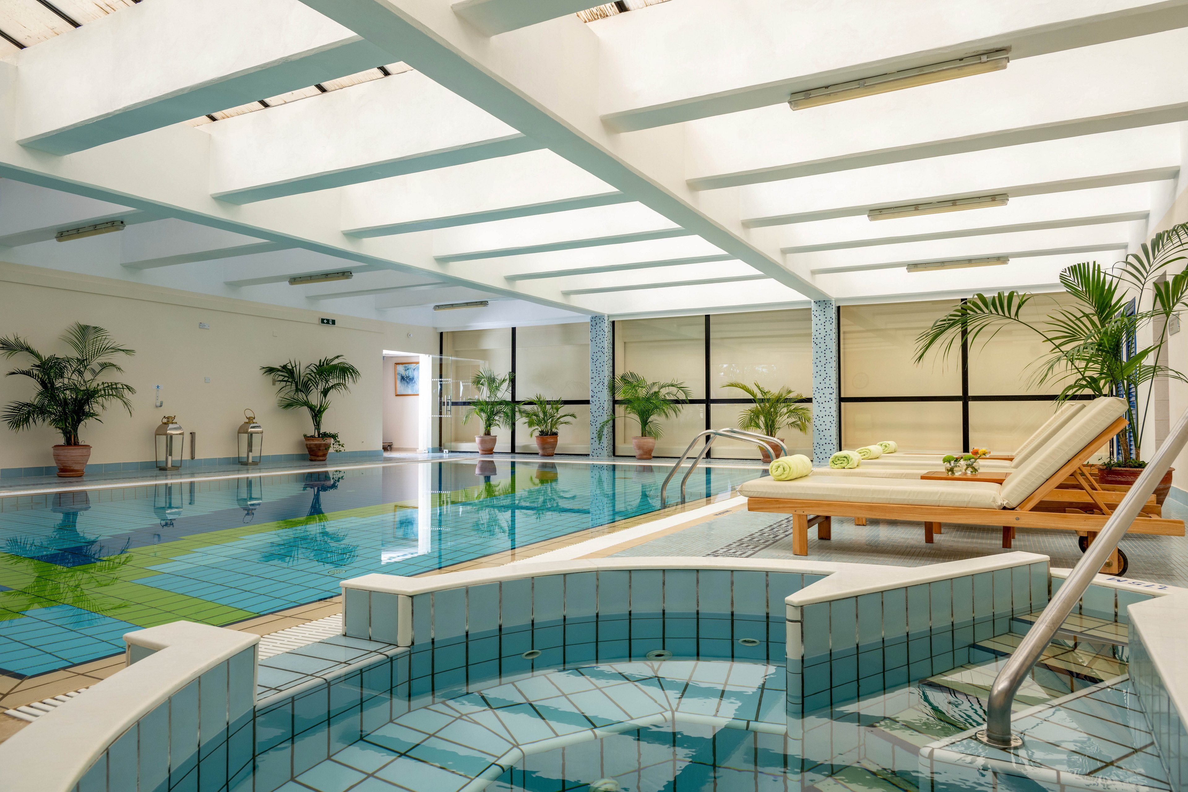 Bright indoor spa area featuring a tiled hot tub in the foreground and a large swimming pool in the background. Wooden lounge chairs with rolled towels sit beside the pool, inviting relaxation. Potted green plants line the walls, adding a natural touch to the clean, modern space. Soft, diffused ceiling light creates a calm and tranquil atmosphere.