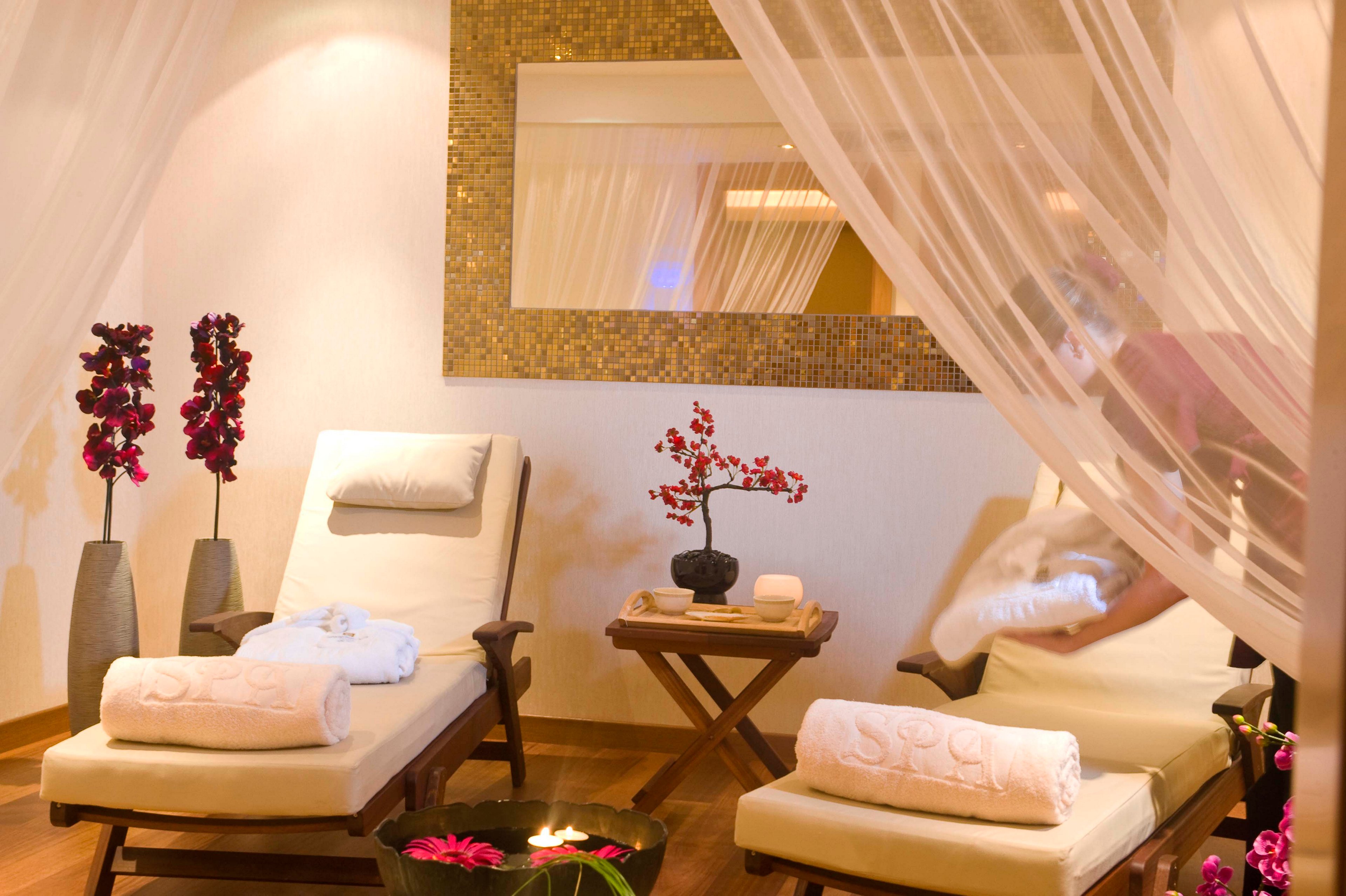 A tranquil spa relaxation room features two cushioned loungers with rolled towels and soft pillows. Sheer white curtains frame the space, while a staff member arranges linens to prepare the area. Decorative elements include tall vases with purple flowers, a small table with a bonsai-style plant, and a bowl of floating candles and petals. Warm lighting and a mosaic-framed wall mirror create a cozy, serene atmosphere.