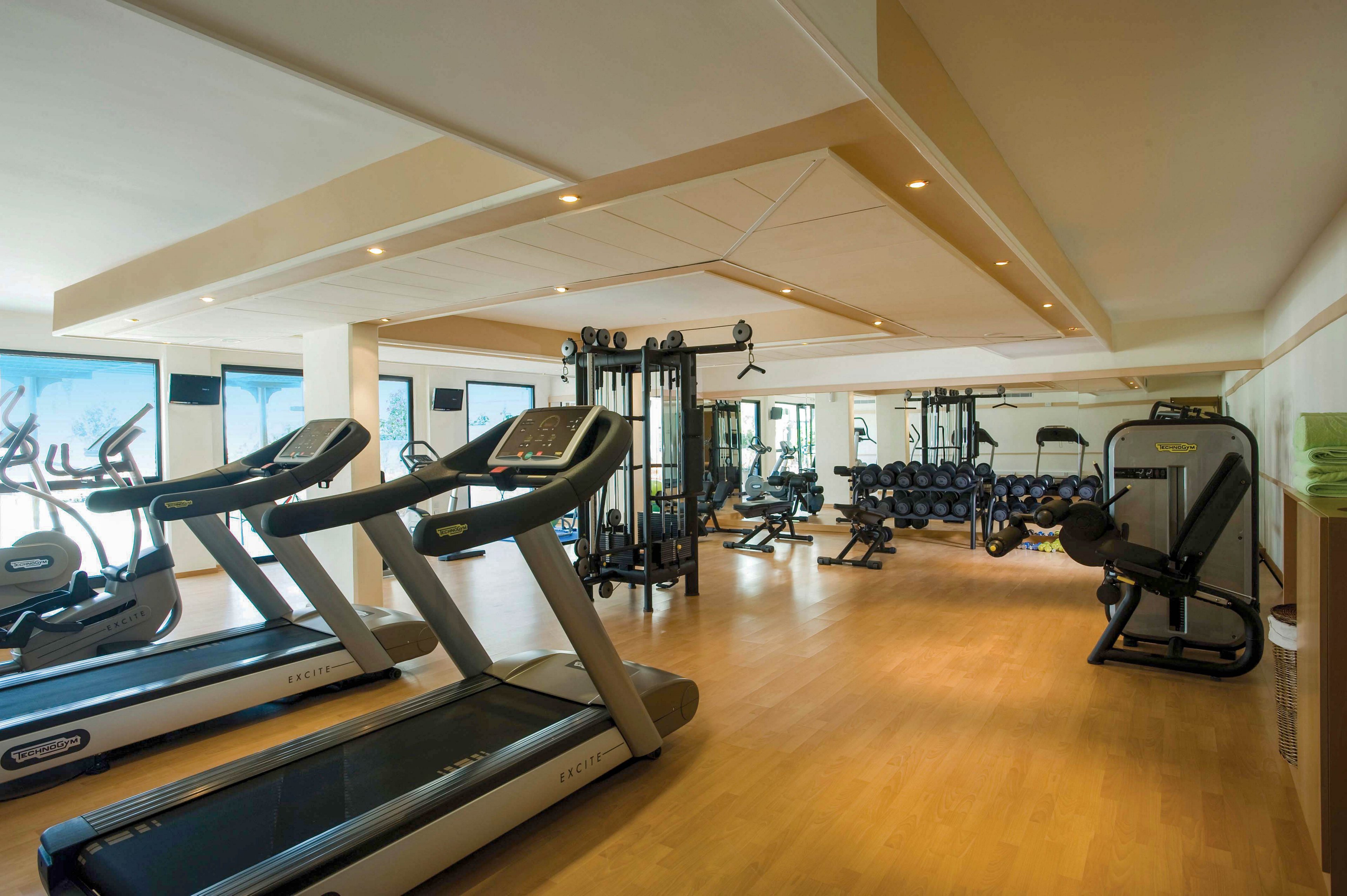 Spacious indoor gym featuring multiple treadmills in the foreground. The room includes weight machines, free weights, and exercise bikes arranged along mirrored walls. Warm wooden flooring and recessed ceiling lights create a clean, inviting workout environment. Large windows provide natural light and views outside, enhancing the contemporary fitness space.