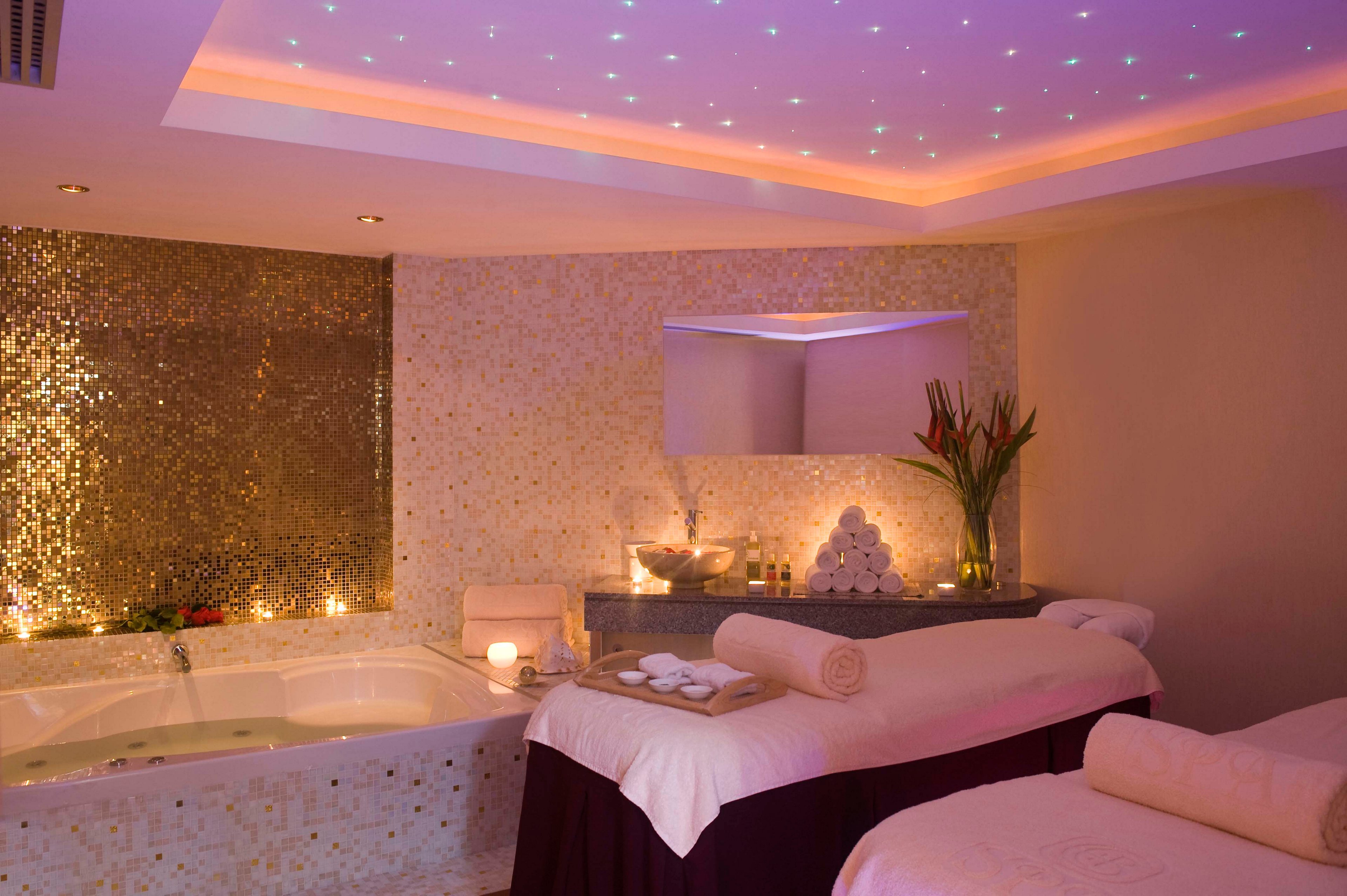 A serene spa treatment room features two massage tables with neatly rolled towels and soft linens. In the corner, a large jacuzzi tub is surrounded by shimmering mosaic tiles and glowing candlelight. The ceiling is lit with soft recessed lighting and tiny star-like points, creating a calming ambiance. A vase of tropical flowers and carefully arranged towels add a touch of elegance and relaxation to the space.