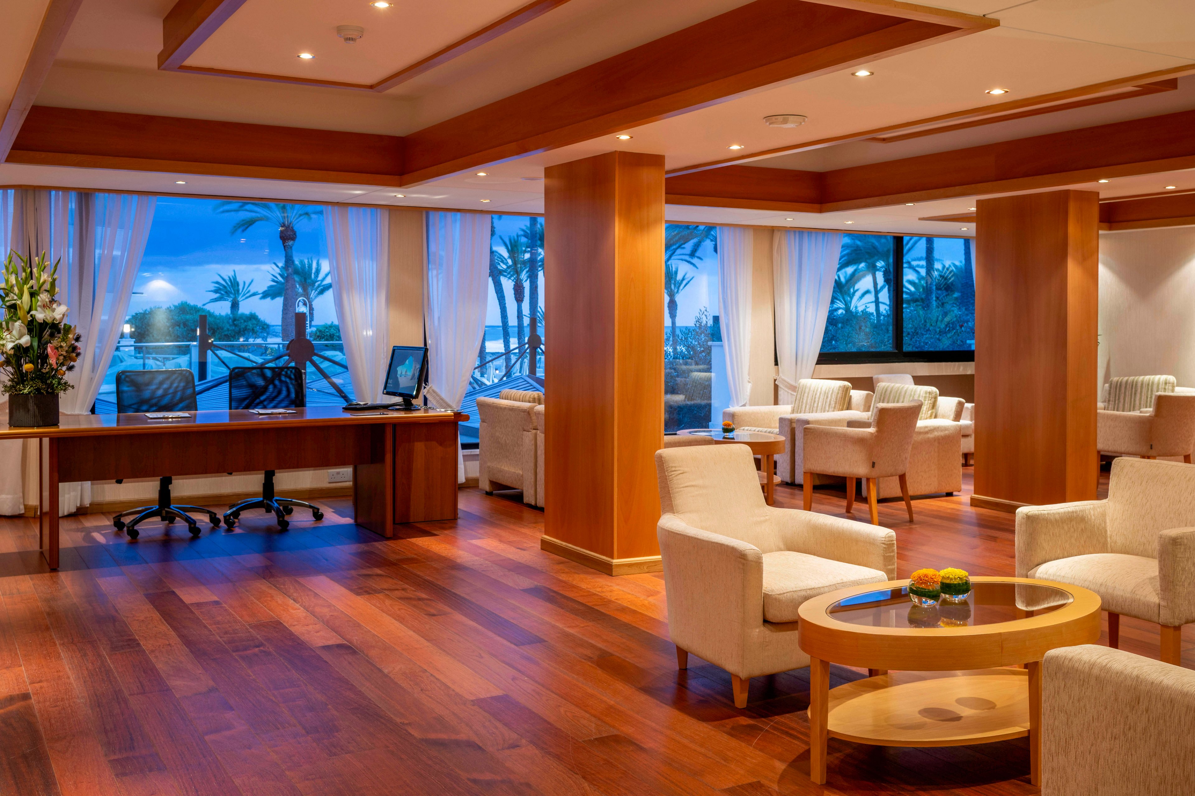 Contemporary hotel lounge area features warm wooden floors and columns with soft upholstered armchairs and round tables. Large panoramic windows reveal palm trees and a serene ocean view at dusk, casting a blue glow into the room. A small desk with a computer and floral arrangement sits near the windows, adding a functional touch to the relaxing space. Overall, the setting feels calm, inviting, and ideal for quiet conversation or unwinding.