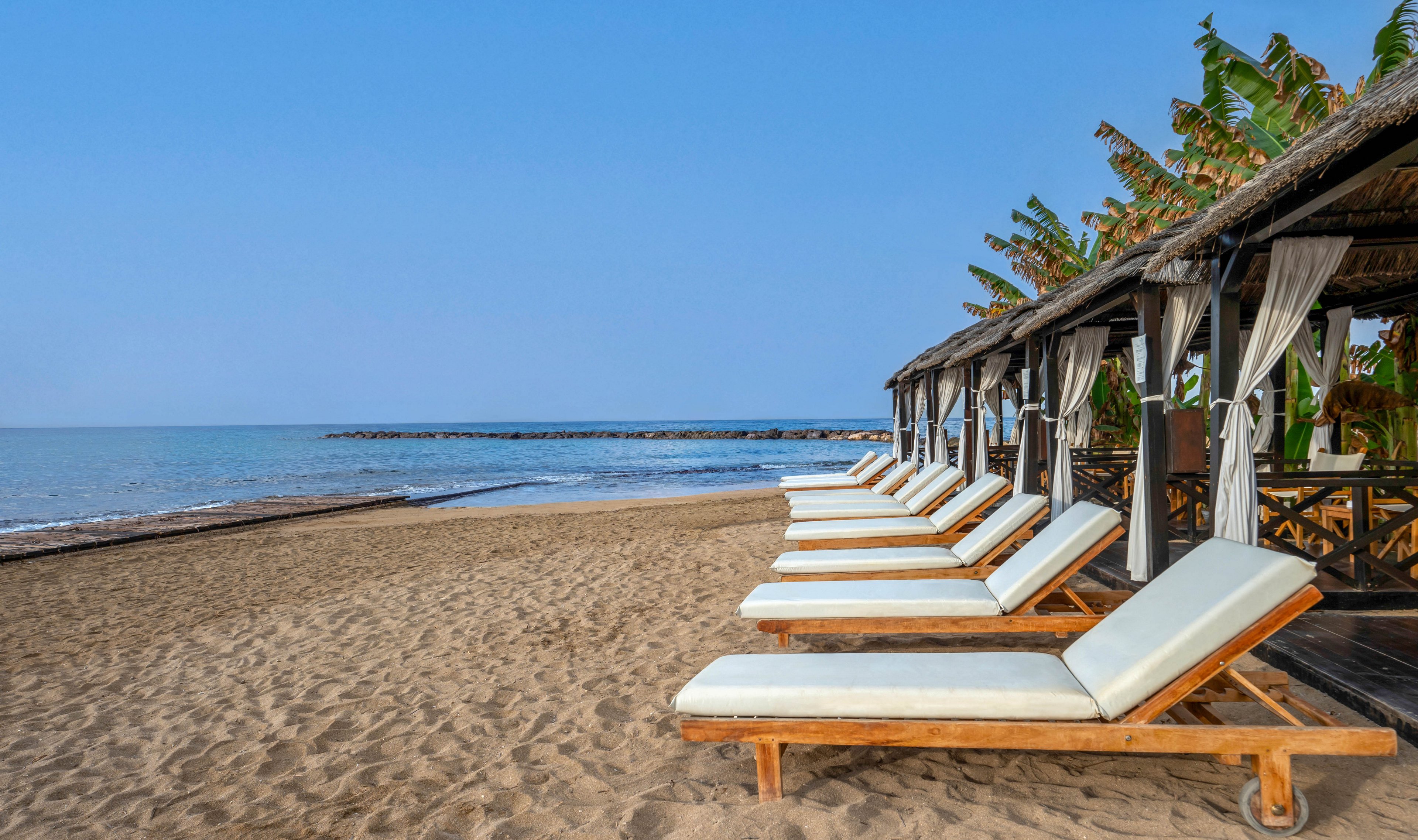 A row of cushioned wooden sun loungers lines a sandy beach beside calm blue water. Thatched cabanas with flowing curtains provide shaded relaxation areas along the shoreline. The scene is quiet and empty, suggesting an early morning or off-peak moment. Palm-like greenery and clear skies contribute to a serene, tropical vacation atmosphere.