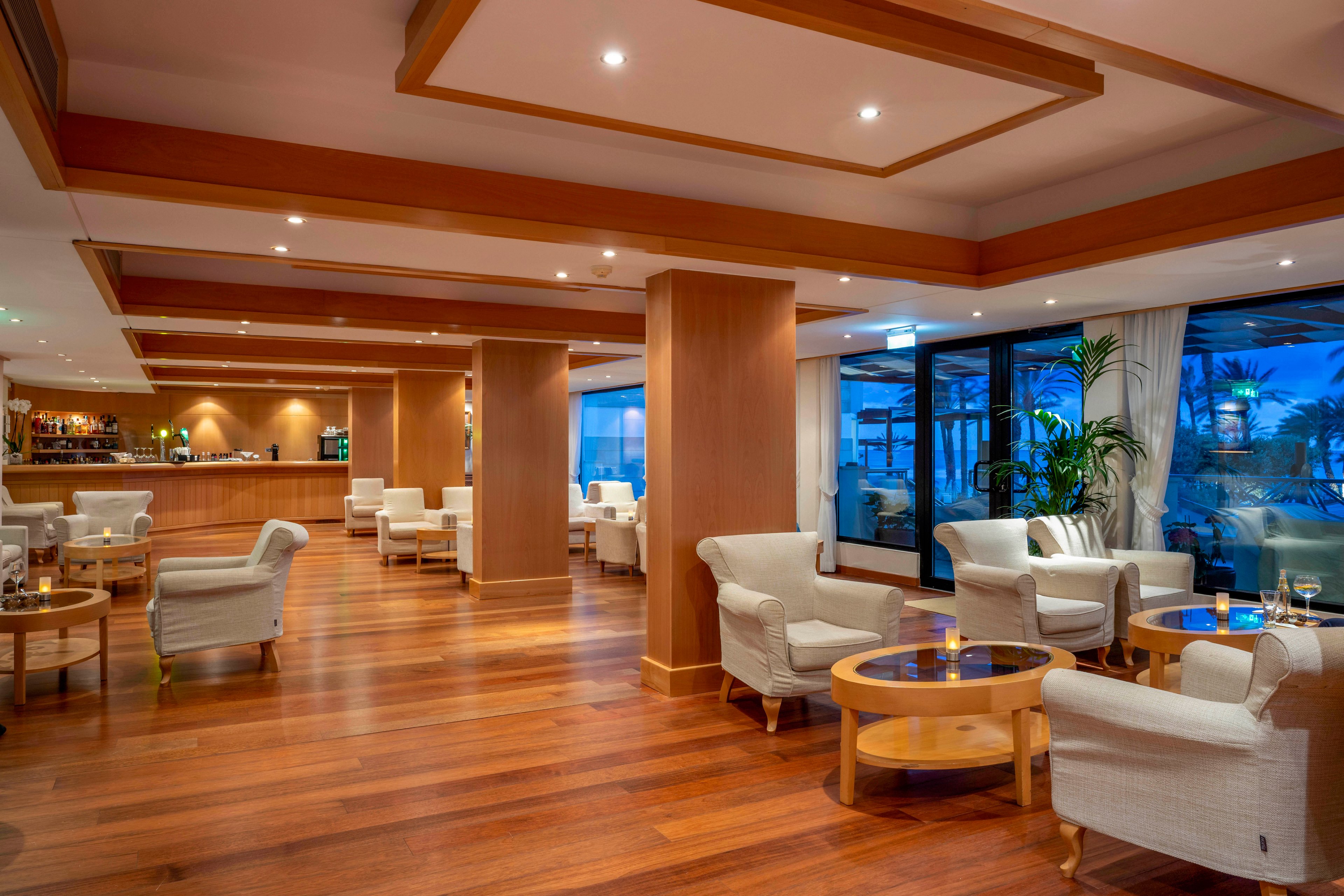 Spacious hotel lounge featuring warm wooden floors and columns is arranged with white upholstered armchairs and round wooden tables. A well-lit bar area is visible in the background, while large windows reveal an evening view of palm trees and blue sky. Soft ambient lighting and neatly set tables with drinks create a relaxed, upscale atmosphere. The space appears designed for socializing, casual meetings, and quiet relaxation.