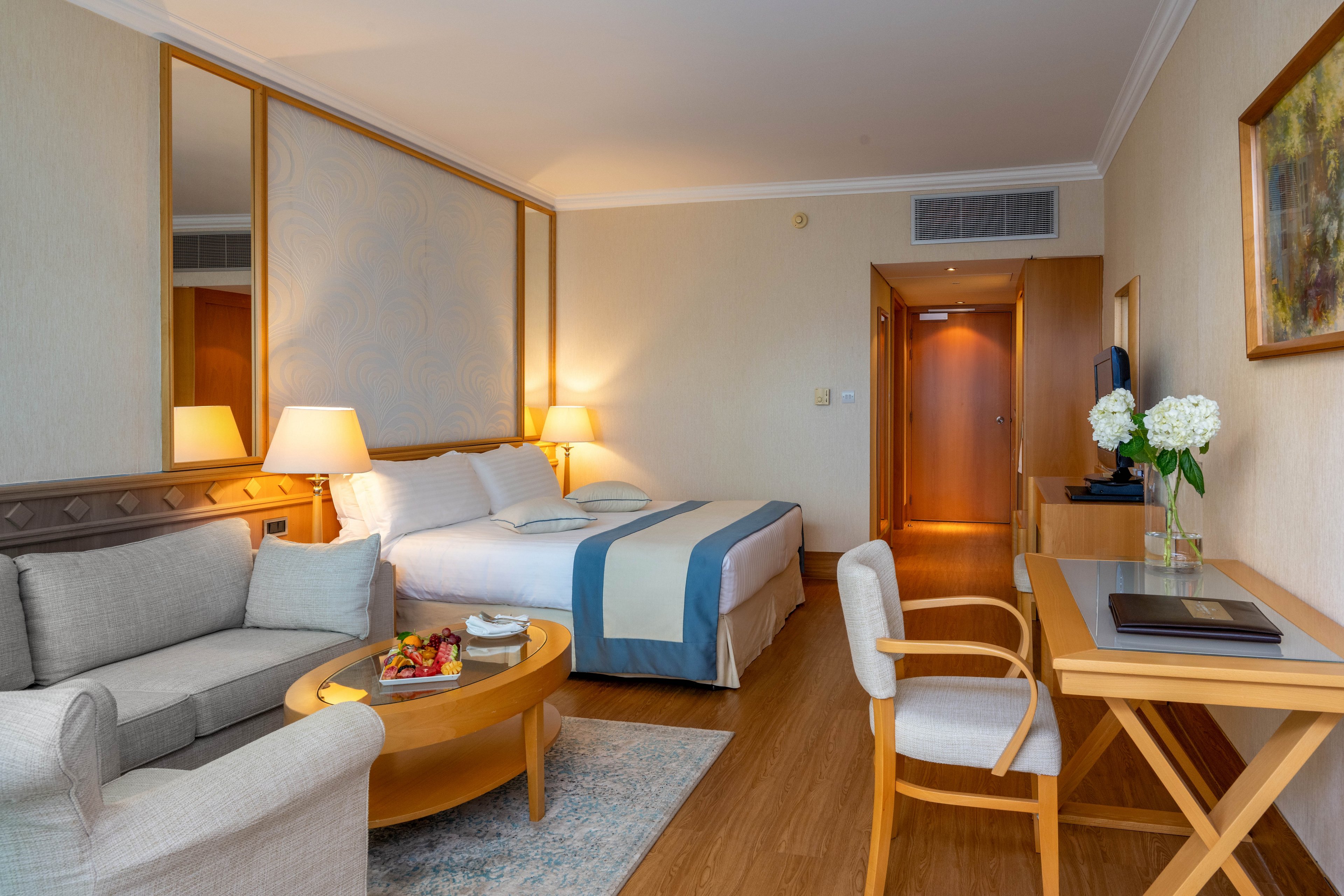 A neatly arranged hotel room features a double bed with blue and beige bedding and soft pillows. Beside the bed, warm table lamps illuminate a small sitting area with a sofa and round wooden coffee table holding a fruit plate. A wooden desk with a chair, laptop, and vase of white flowers creates a functional workspace near the wall. The room has light-colored walls, wooden flooring, and a doorway leading to the entrance, conveying a clean and inviting atmosphere.
