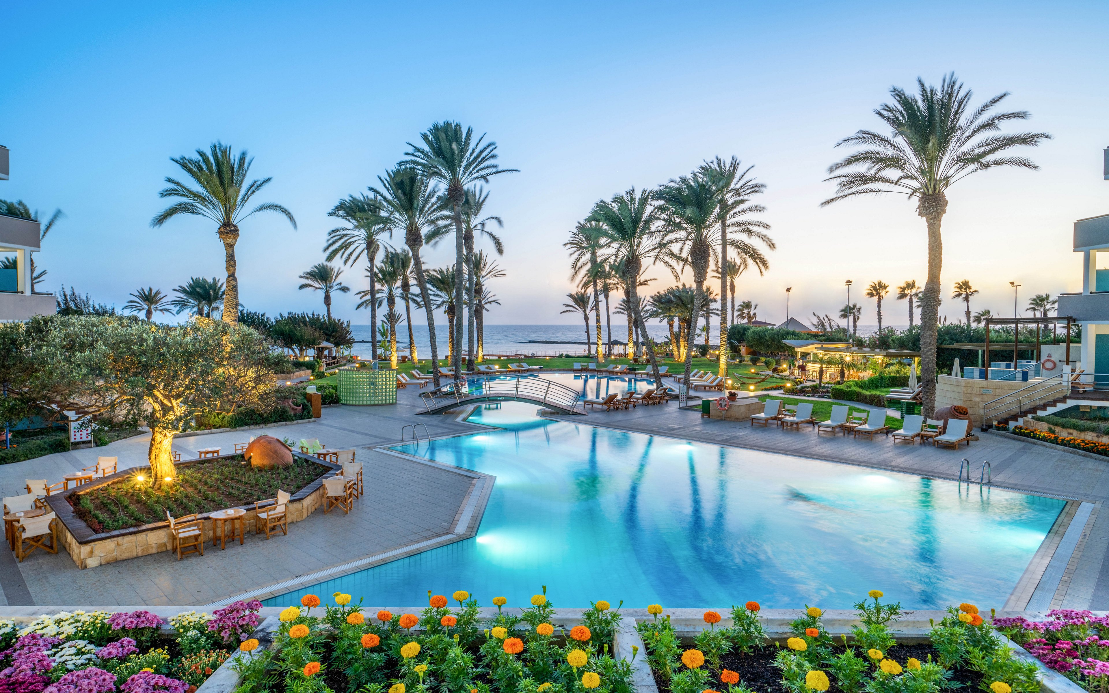 A large, inviting resort swimming pool is surrounded by sun loungers and landscaped gardens filled with colorful flowers. Tall palm trees frame the scene and lead the eye toward the ocean in the background at dusk. Modern buildings and outdoor seating areas with wooden tables and chairs create a relaxed vacation atmosphere. The calm water of the pool reflects the soft evening sky, enhancing the tranquil, tropical setting.
