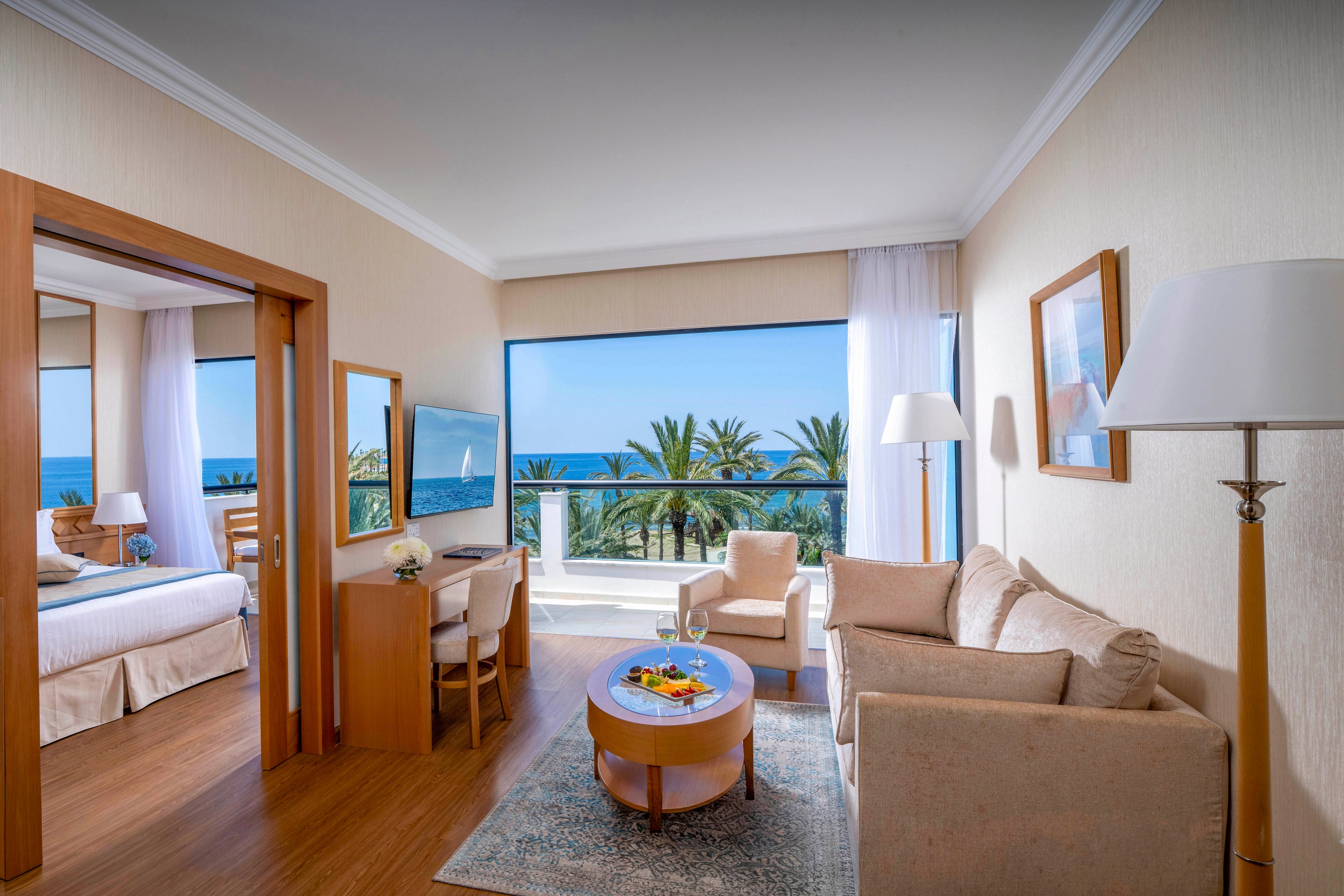 Spacious hotel suite features a cozy living area with neutral-toned furniture and a separate bedroom. Large windows and a balcony showcase a scenic ocean view framed by palm trees. A small dining table, floor lamp, and wall art add to the contemporary decor. Natural light fills the room, creating a relaxing and inviting vacation atmosphere.