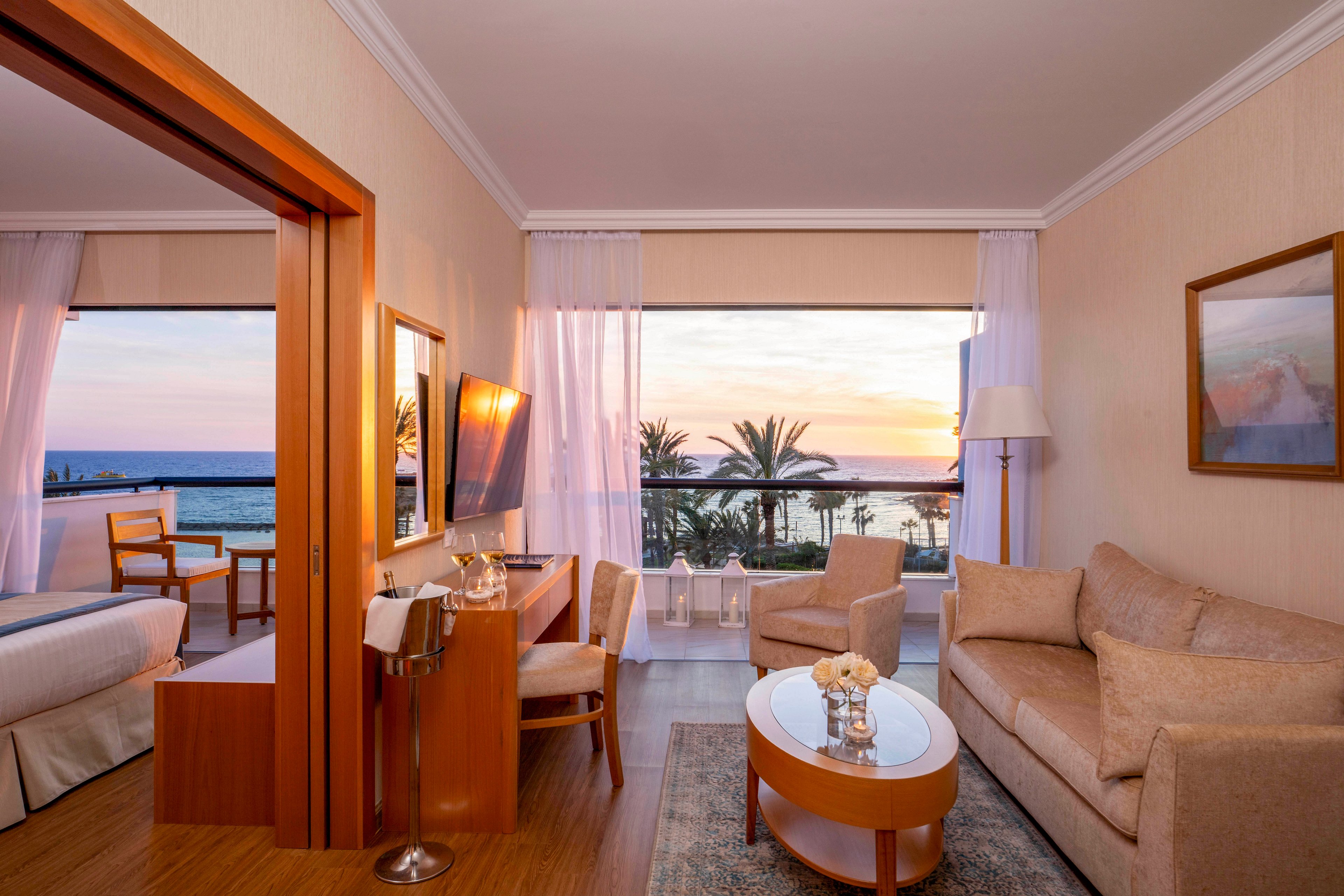 Elegant hotel suite featuring a cozy living area with sofa, armchair, and coffee table. A separate bedroom with a bed and wooden chair opens toward large windows overlooking the ocean. Sheer white curtains frame the sunset sky and palm trees outside, filling the room with warm natural light. Soft neutral tones and contemporary furnishings create a relaxing, upscale atmosphere.
