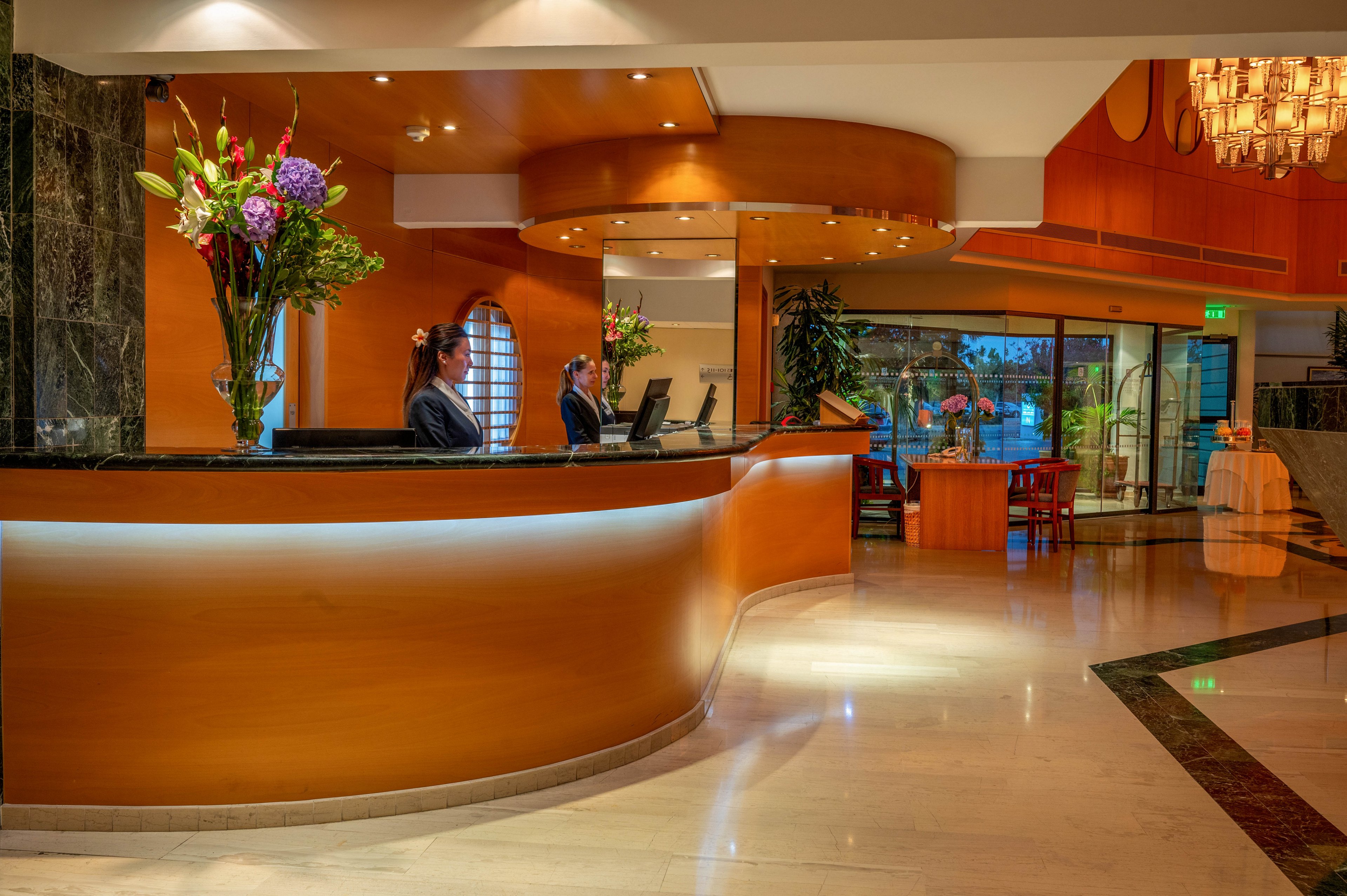 A modern hotel reception area features a curved wooden front desk with two receptionists working at computers. The lobby is warmly lit, highlighting polished marble floors and contemporary decor. Large floral arrangements add color near the desk, while glass doors and seating are visible in the background. The overall atmosphere is professional, welcoming, and upscale.