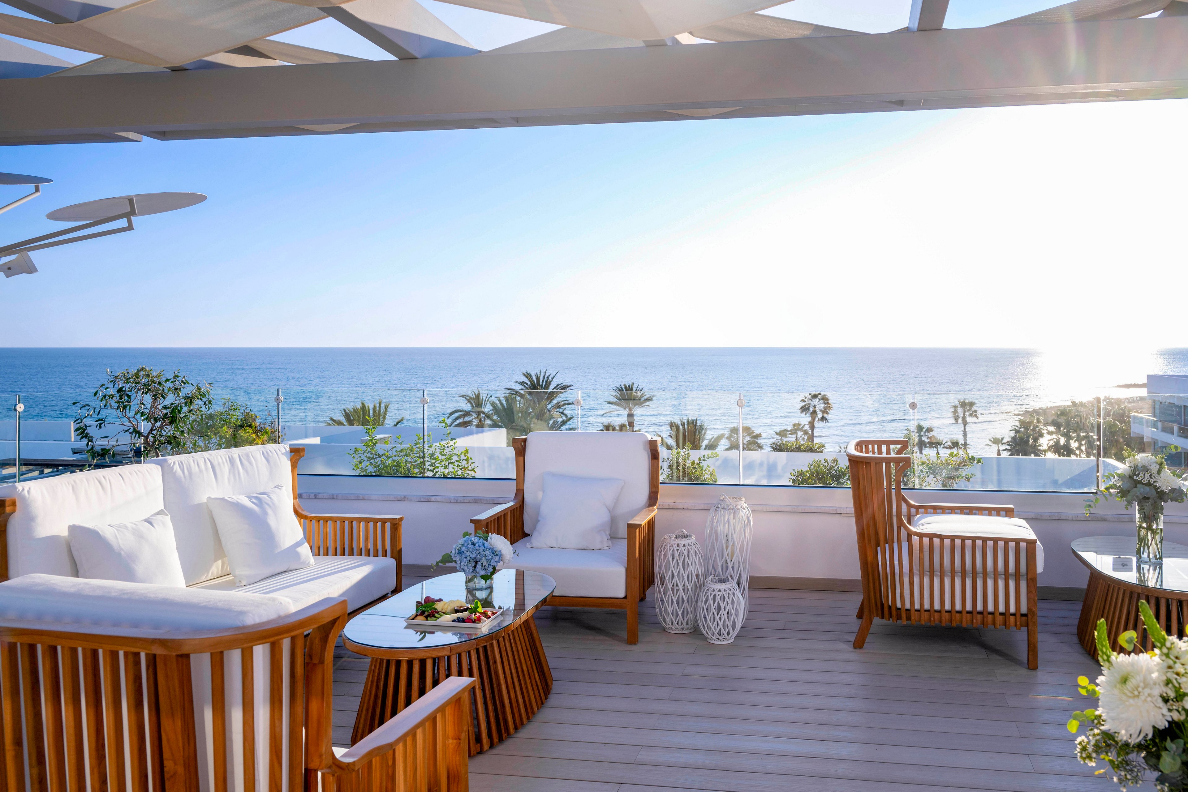 A woman in a long white dress stands on a stylish terrace overlooking the ocean. Contemporary wooden lounge furniture with white cushions is arranged around small tables decorated with flowers. The bright sunlight reflects off the sea, creating a serene and luxurious atmosphere. Palm trees and coastal buildings are visible in the distance, enhancing the resort-like setting.