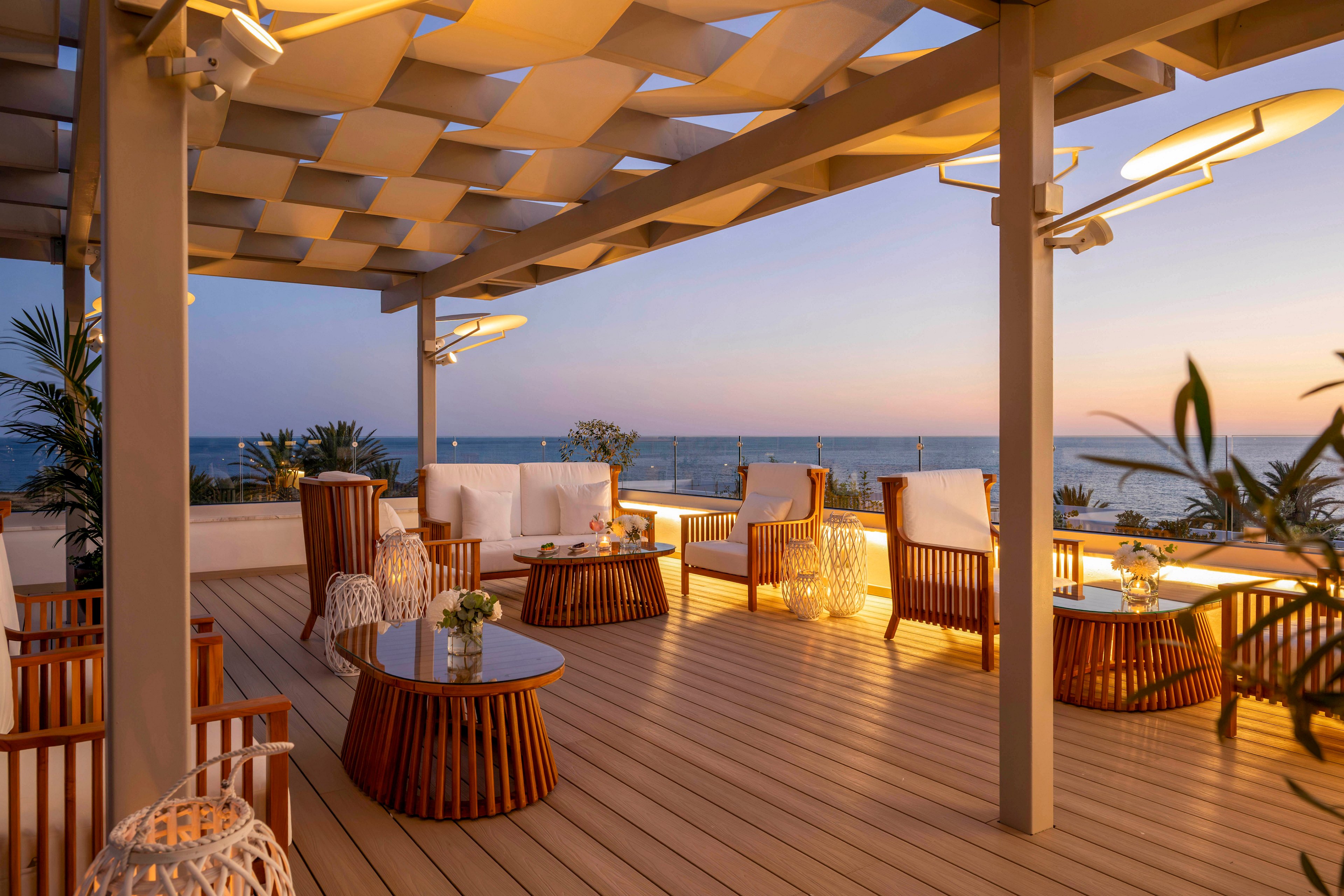 An elegant outdoor terrace lounge overlooks the sea at sunset. Wooden lounge chairs and sofas with white cushions are arranged around glass-topped tables under a modern pergola. Warm ambient lighting and decorative lanterns create a cozy, inviting atmosphere. The calm ocean and pastel evening sky provide a serene backdrop for relaxation or social gatherings.