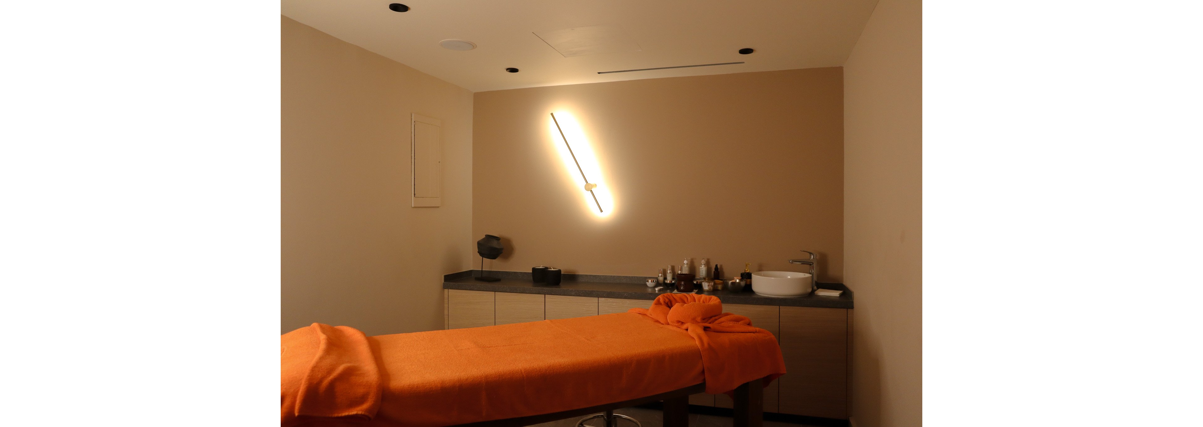 A softly lit spa treatment room features a massage table covered with an orange towel. The space has warm beige walls and a modern diagonal wall light creating a relaxing glow. Along the back wall, a countertop holds various bottles, jars, and a white basin sink. The overall atmosphere is calm and inviting, ideal for wellness and relaxation.