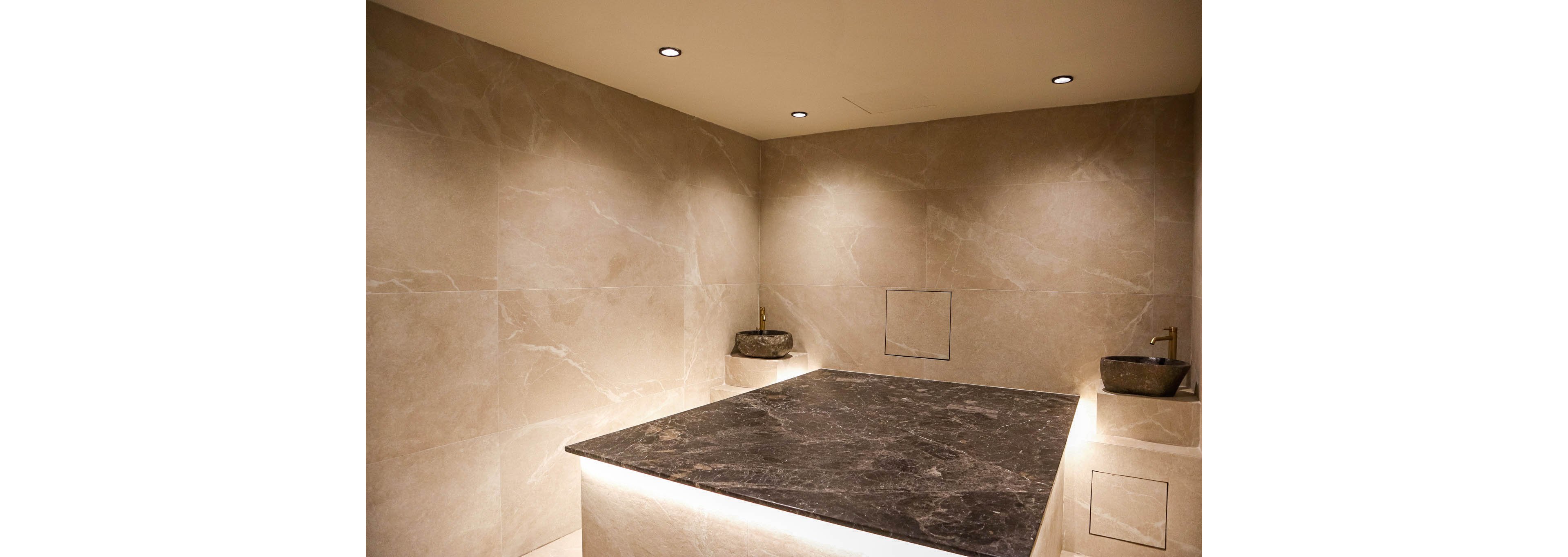 A contemporary steam room features a large dark stone slab in the center, illuminated from below. Smooth beige stone tiles cover the walls and built-in benches, creating a clean, minimalist look. Two small stone basins with wall-mounted taps are positioned on opposite sides of the room. Recessed ceiling spotlights provide warm, focused lighting that enhances the spa-like atmosphere.