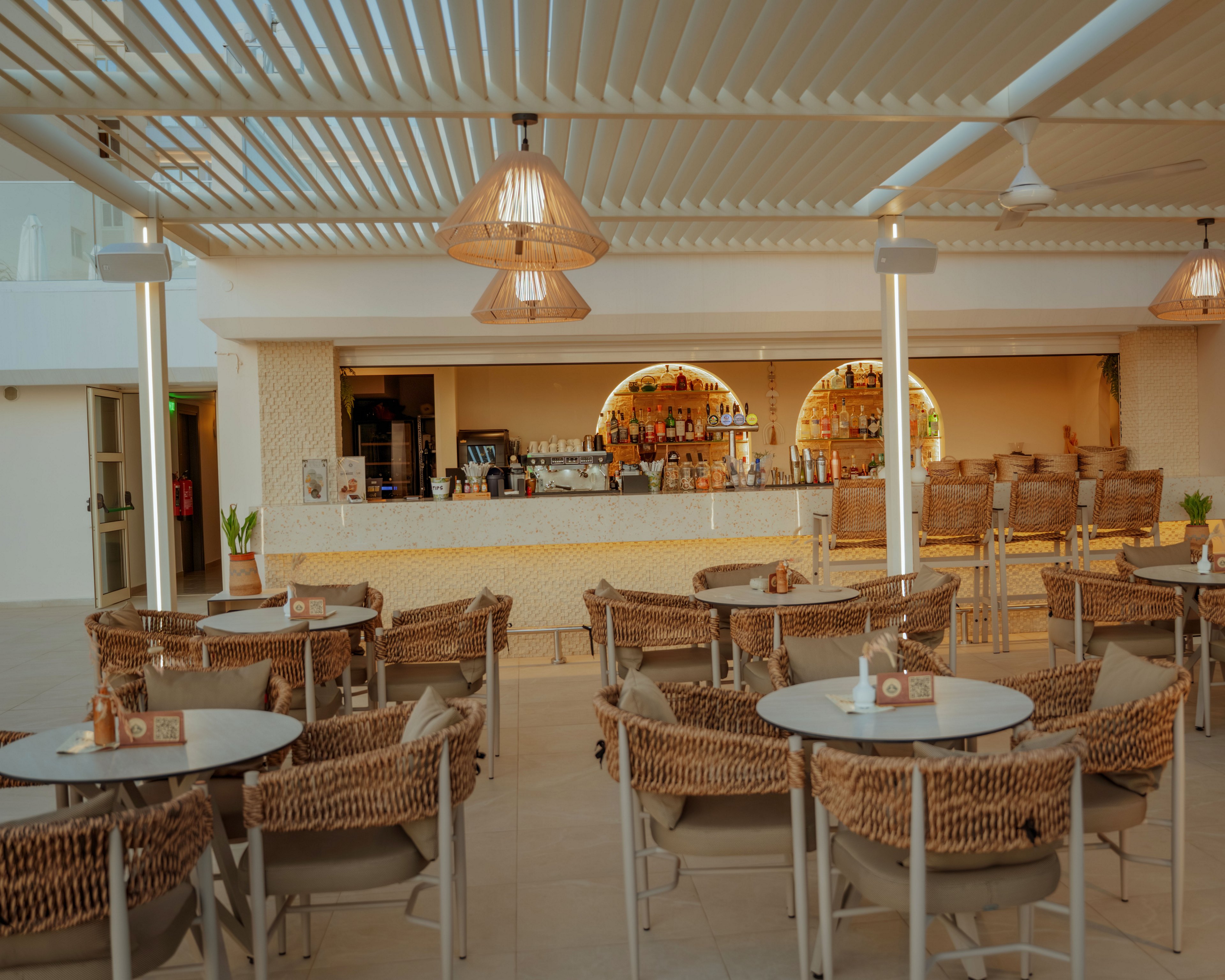 An open-air bar area features rows of round tables surrounded by woven rattan-style chairs. The bar counter in the background is stocked with bottles and glassware, framed by two softly lit archways. Warm pendant lights hang from a slatted pergola ceiling, creating a relaxed, coastal atmosphere. Neutral tones and natural textures give the space a modern, inviting look.