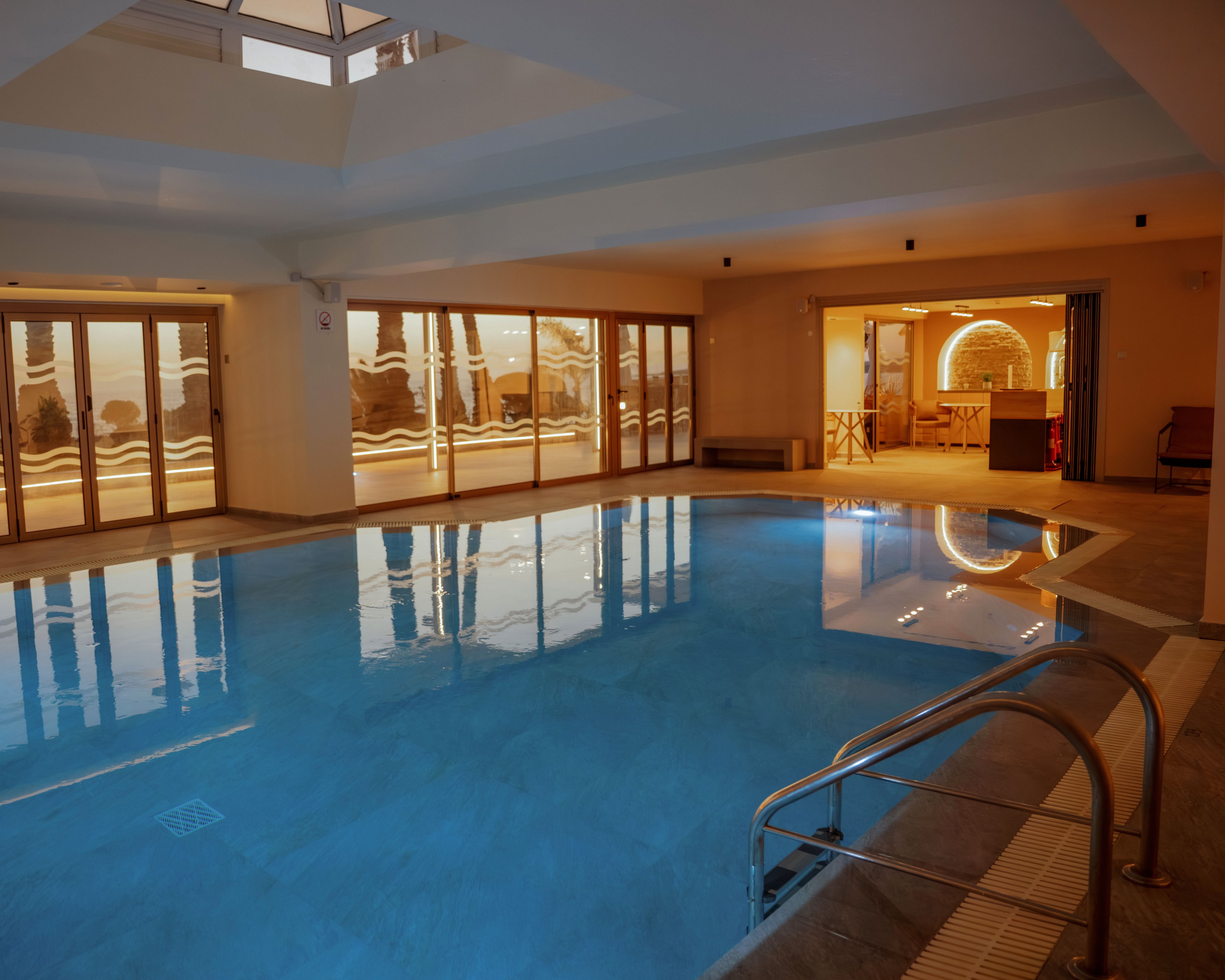 A tranquil indoor swimming pool is illuminated by warm ambient lighting and reflections from large windows. The pool area features metal handrails, tiled flooring, and a calm blue water surface. Beyond the glass doors, a cozy lounge or bar area glows with decorative lights, enhancing the inviting atmosphere. The overall scene suggests a relaxing leisure space in a hotel or spa during evening hours.