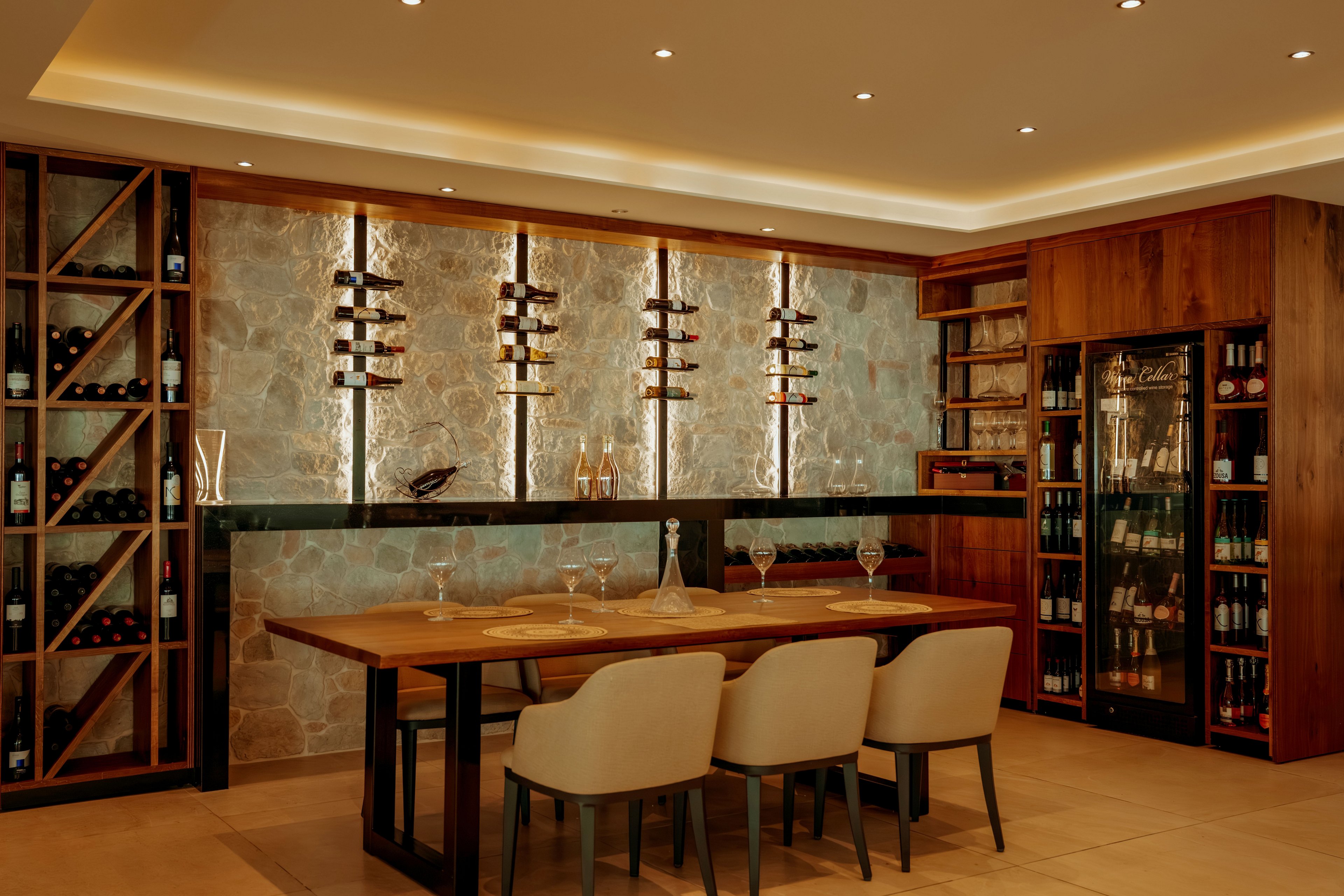 A sophisticated wine tasting room features a long wooden table set with glassware and decanters. Wall-mounted racks display bottles of wine against a textured stone backdrop, highlighted by warm accent lighting. Built-in wooden cabinets and shelves hold additional bottles and glassware, creating a refined cellar atmosphere. Neutral upholstered chairs and recessed ceiling lights contribute to the cozy, upscale ambiance.