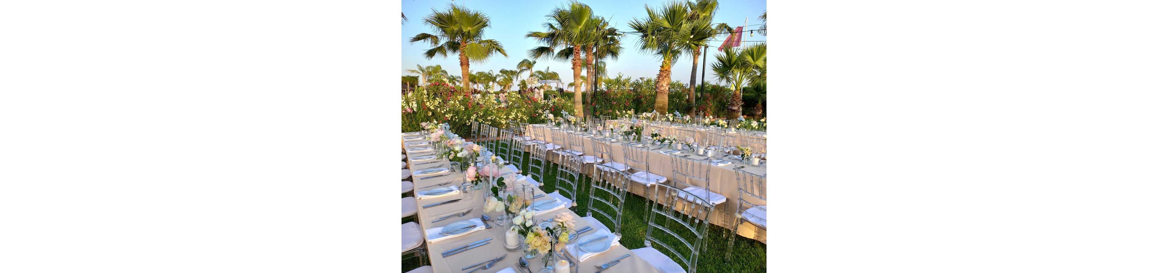 Long banquet tables are arranged outdoors in a lush garden setting lined with palm trees. Transparent chairs and neatly folded napkins create a refined, coordinated look. Floral centerpieces with white and pastel flowers run along the length of the tables. The scene is bathed in warm daylight, suggesting a festive event such as a wedding or celebration.