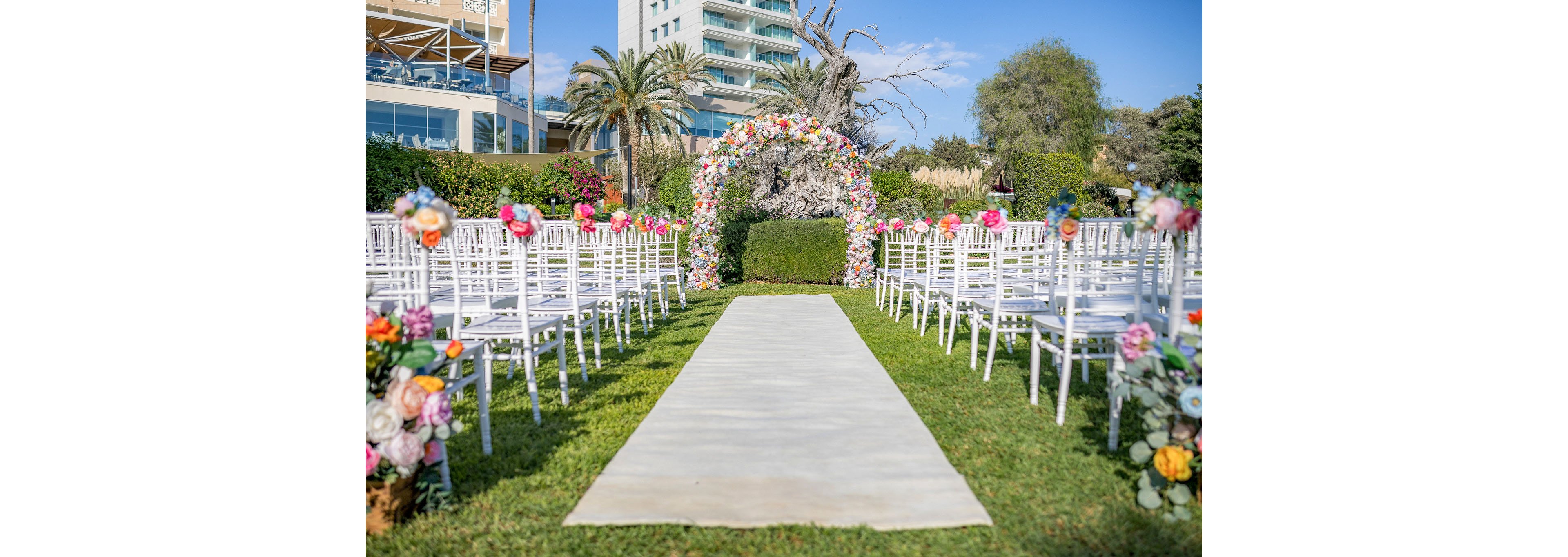 An outdoor wedding ceremony setup features a white aisle runner leading to a lush floral arch. Rows of white chairs line both sides of the aisle, each decorated with colorful flower arrangements. The scene is set on a green lawn with palm trees, manicured hedges, and a modern building in the background. Bright daylight and clear skies create a cheerful, romantic atmosphere.