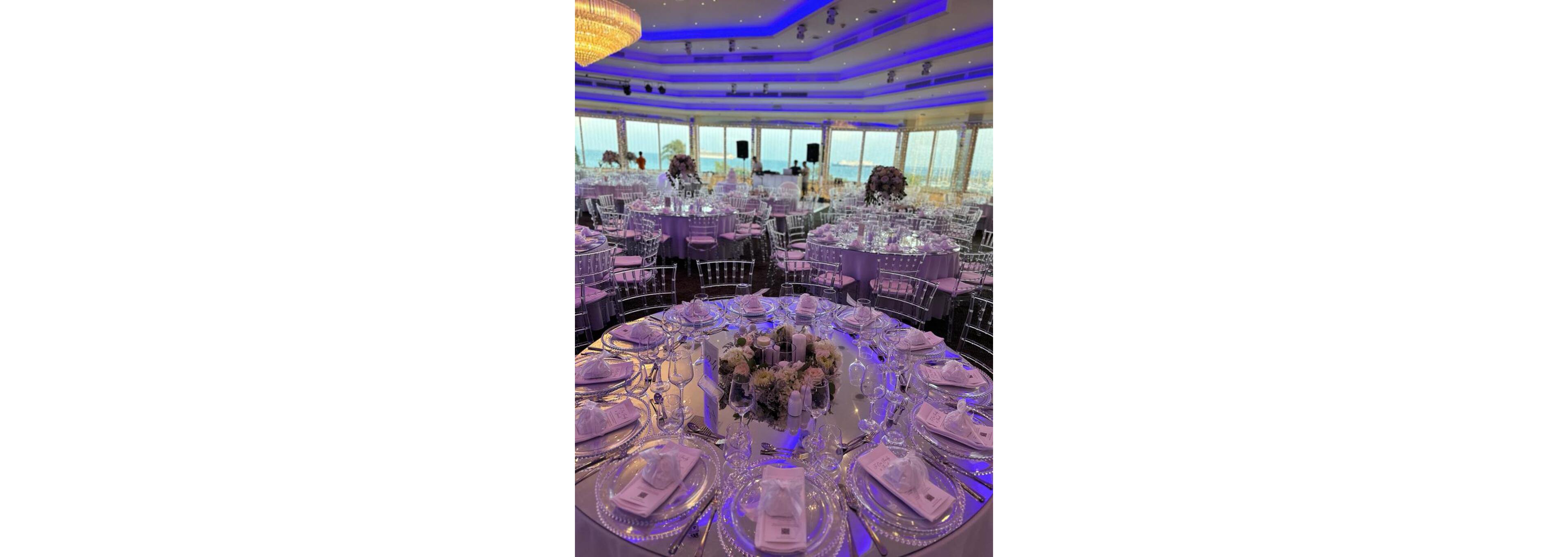A luxurious banquet hall is set up for a formal event with round tables covered in white linens and transparent chairs. Each table features neatly arranged glassware, plates, and folded napkins, centered around floral arrangements and candles. The room is illuminated with purple and blue ambient lighting from the ceiling, creating a sophisticated atmosphere. Large windows reveal a scenic waterfront view in the background, enhancing the upscale setting.