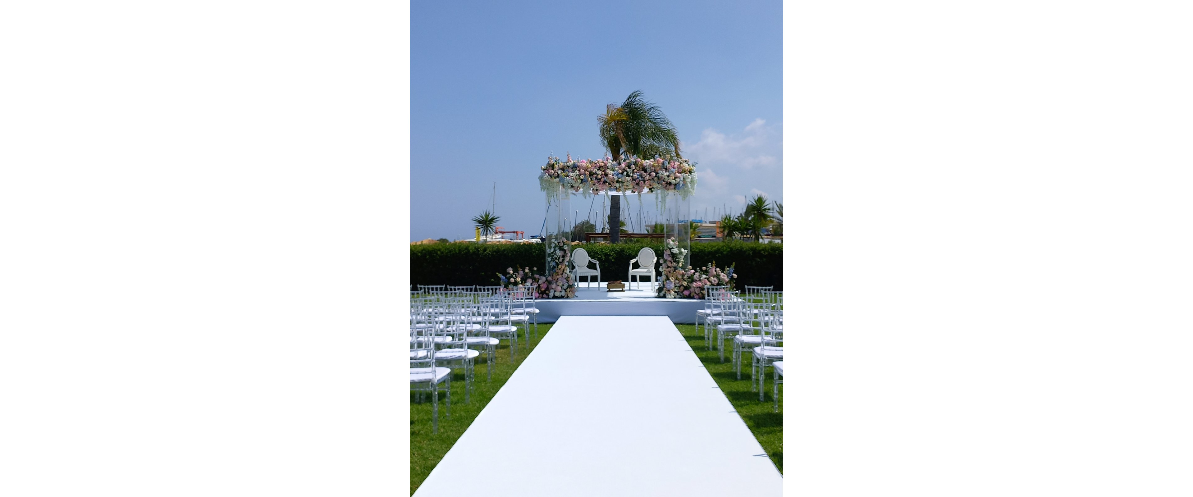 An outdoor wedding ceremony setup features a long white aisle leading to a raised stage. Two white chairs sit beneath a floral arch adorned with pastel flowers and greenery. Transparent chairs are arranged in neat rows on either side of the aisle over a green lawn. A tall palm tree rises behind the arch, with a clear blue sky in the background. The scene suggests a bright, elegant celebration in a warm, coastal setting.