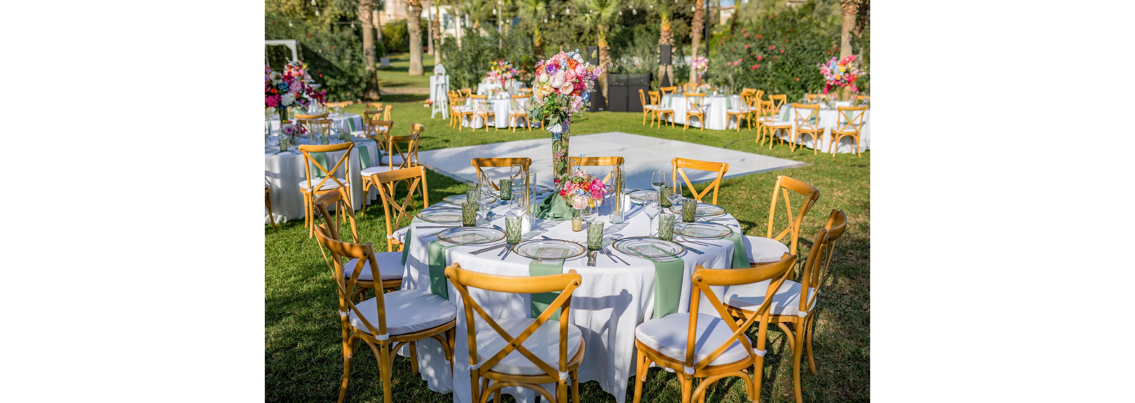 A round table is set for an outdoor event in a lush garden. White linens with soft green runners are paired with wooden cross-back chairs and clear glass plates. A tall floral centerpiece with pink, orange, and purple flowers decorates the middle of the table. Additional similarly decorated tables and greenery are visible in the background, suggesting a festive celebration or wedding reception.