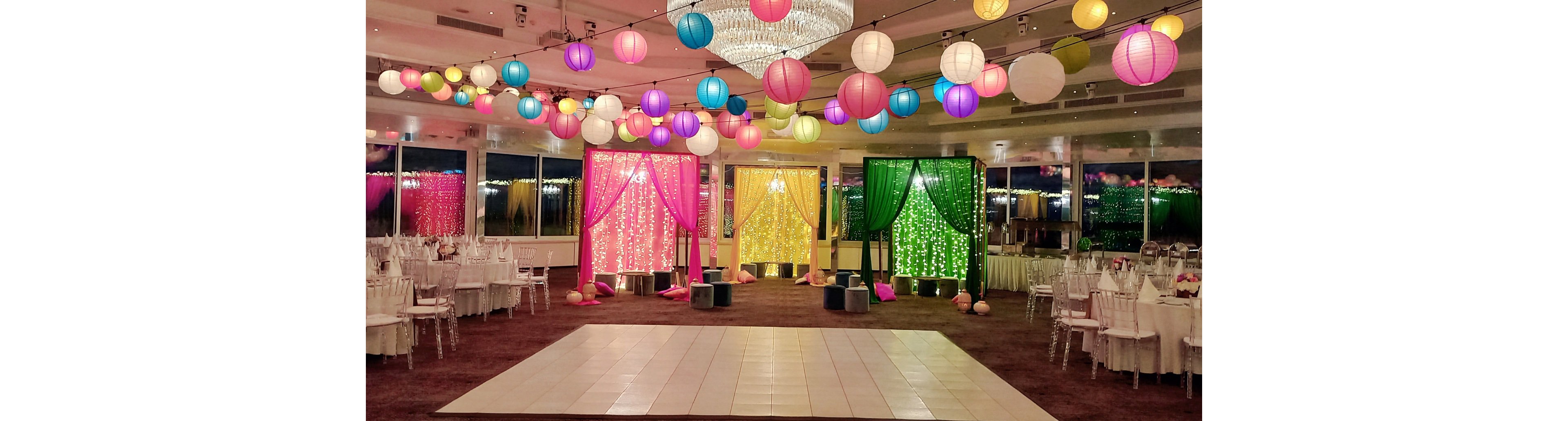 A large banquet hall is decorated with colorful hanging paper lanterns and illuminated draped backdrops. Pink, yellow, and green curtain panels with string lights frame the far wall, creating a vibrant focal point. Round tables with white linens and chairs surround an empty central dance floor. The room is set for a festive celebration or wedding reception with bright, playful lighting.