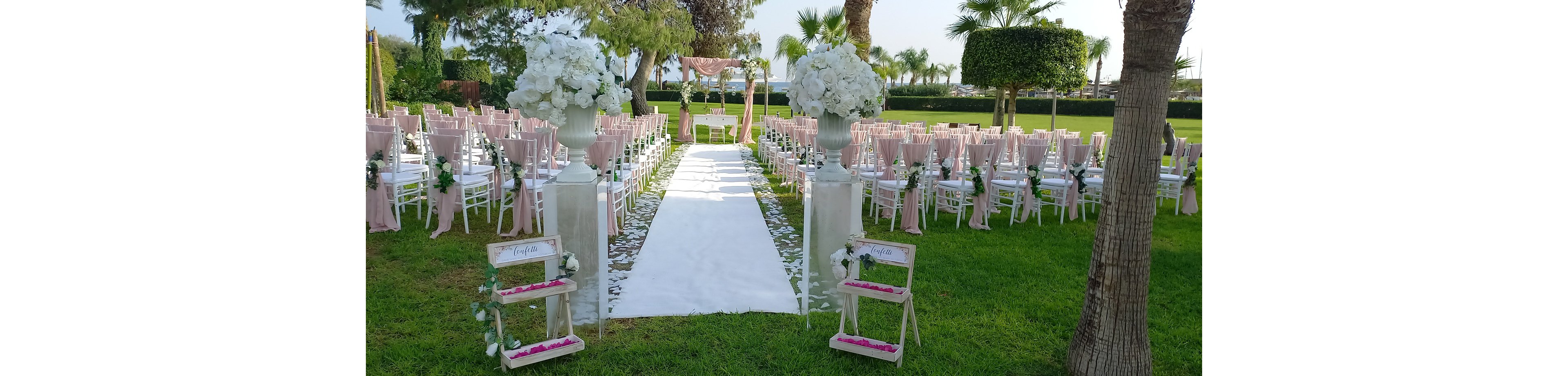 An elegant outdoor wedding ceremony is arranged on a manicured lawn with a long white aisle runner leading to a floral arch. Rows of white chairs with soft pink accents line both sides of the aisle, creating a symmetrical and romantic setting. Tall white flower arrangements stand at the entrance of the aisle, complemented by small decorative stands with petals. Palm trees and trimmed hedges frame the background, enhancing the serene garden atmosphere.