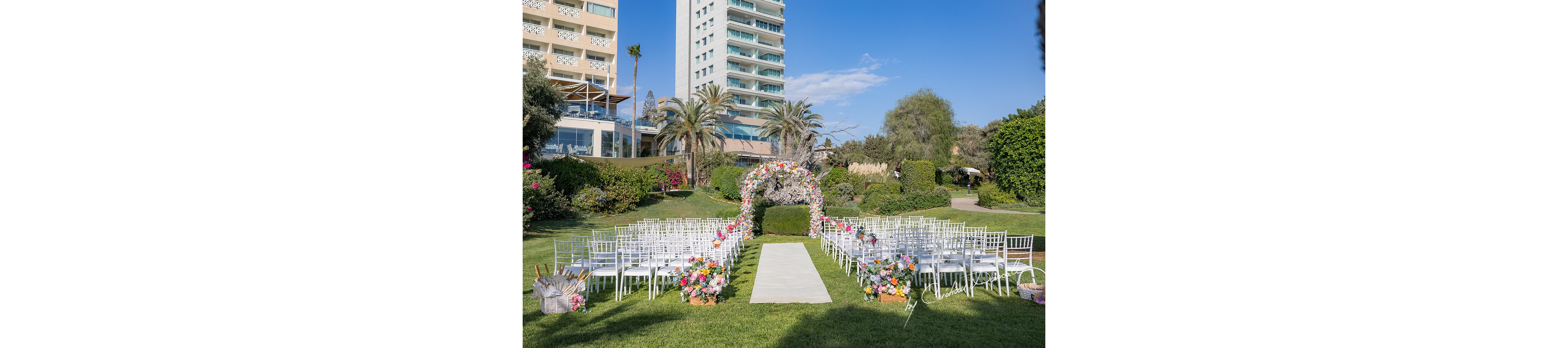 An elegant outdoor wedding ceremony is arranged on a manicured lawn in front of a floral arch. White chairs are lined in rows on both sides of a central aisle decorated with colorful flower arrangements. Tall palm trees, lush greenery, and landscaped bushes surround the venue, with modern hotel buildings visible in the background. The bright blue sky and sunlight create a cheerful, romantic atmosphere.